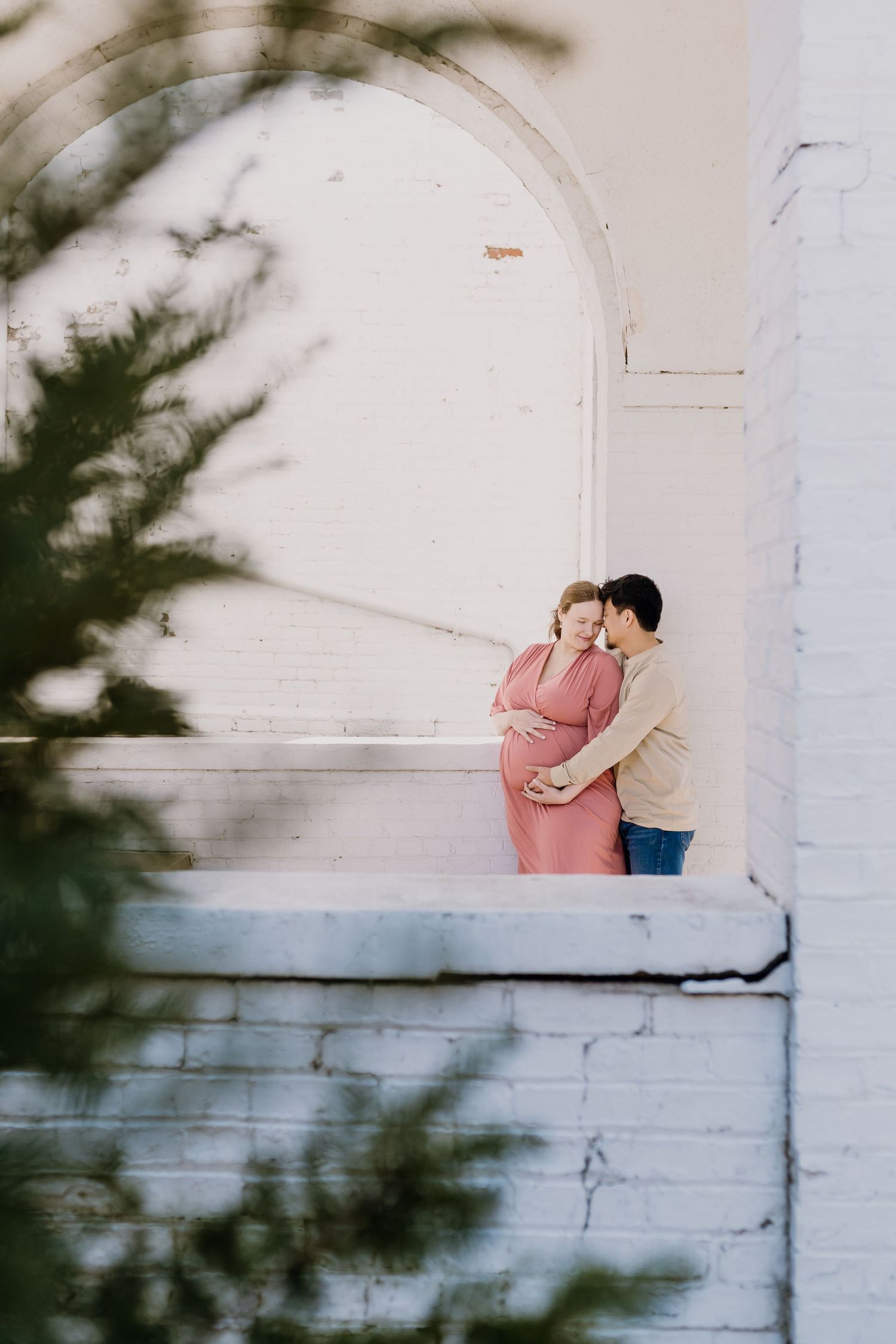Pregnant woman holds belly and is embraced by husband as she looks down smiling, framed by a historic building's arch