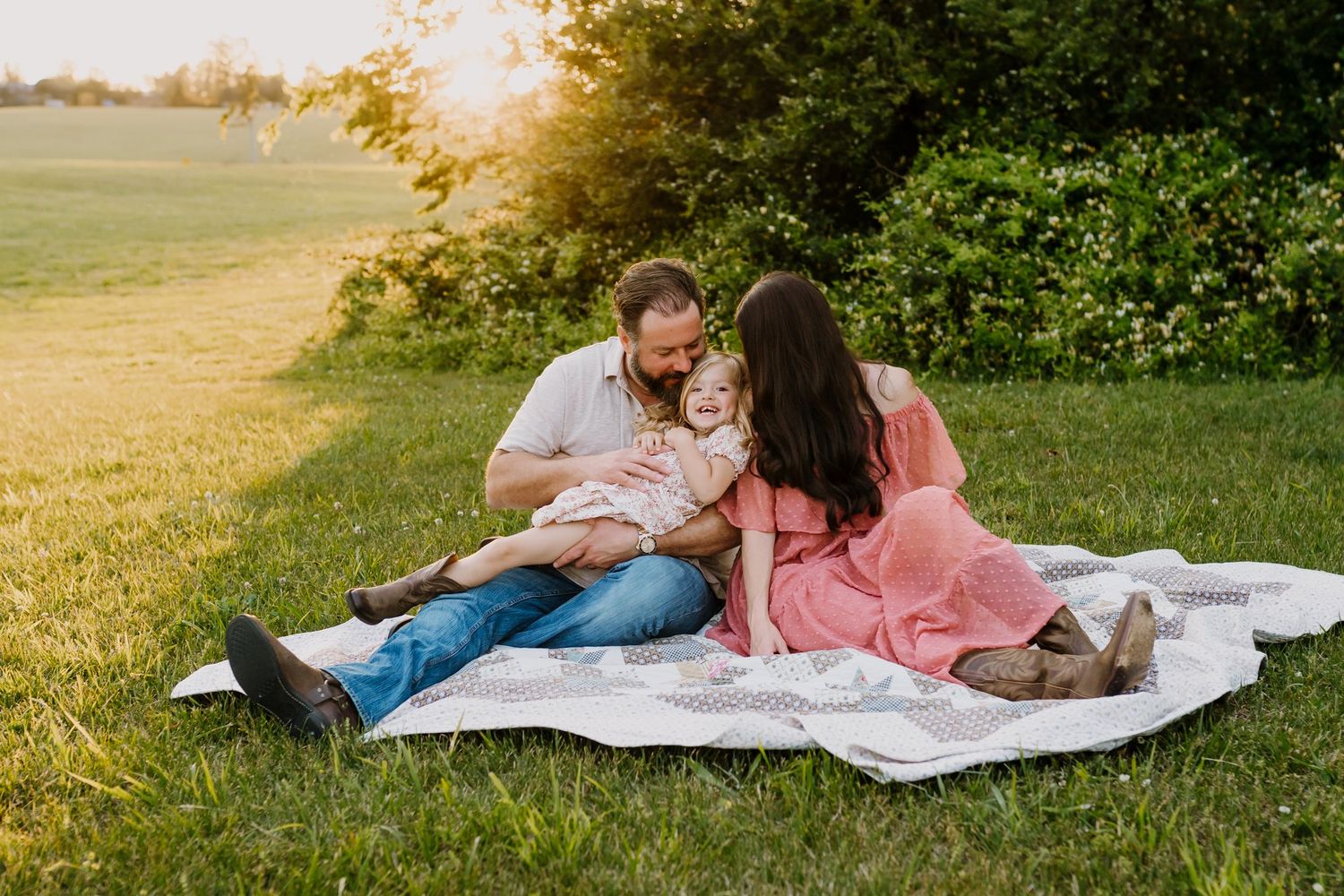 Family of three sitting on a blanket in a sunny green field at golden hour.