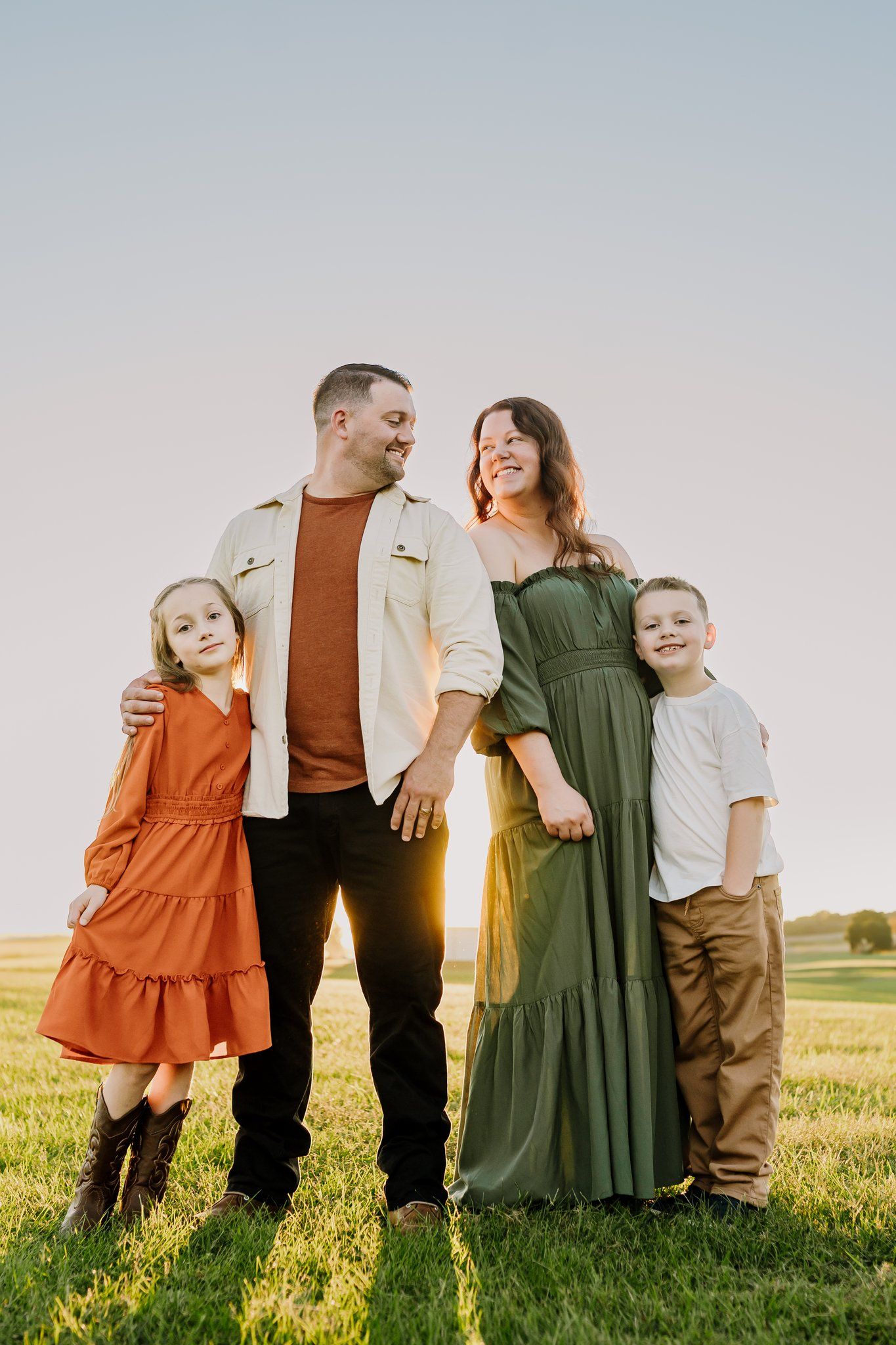 Family of four posing in a sunlit green field at golden hour, parents smiling at each other with children by their sides.