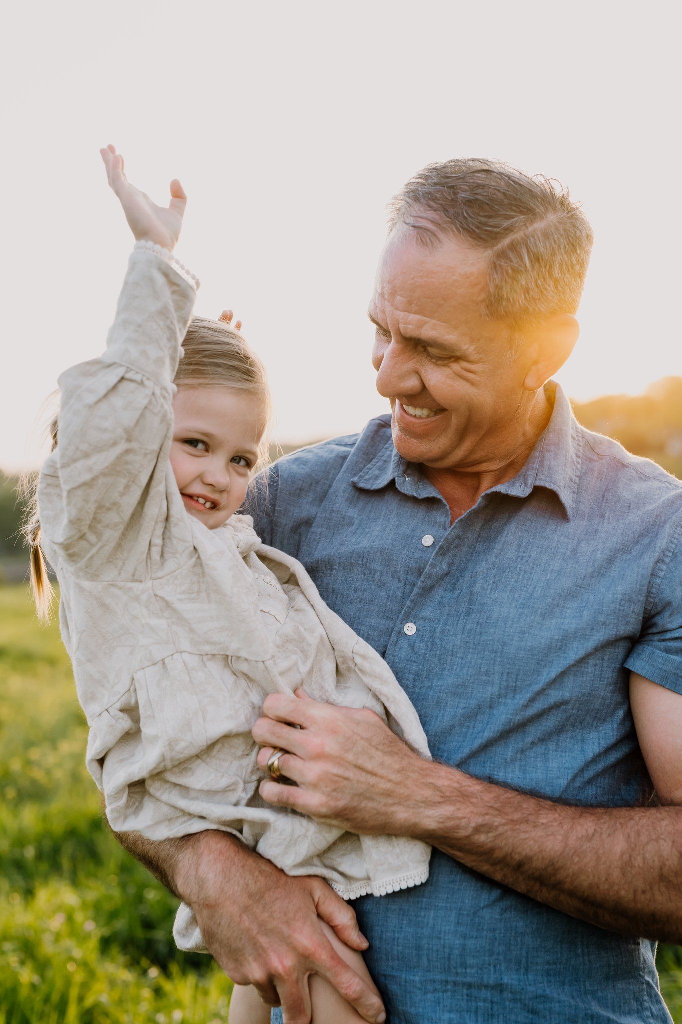 Smiling grandfather holding cheerful young girl with raised arm in sunny outdoor field at golden hour.