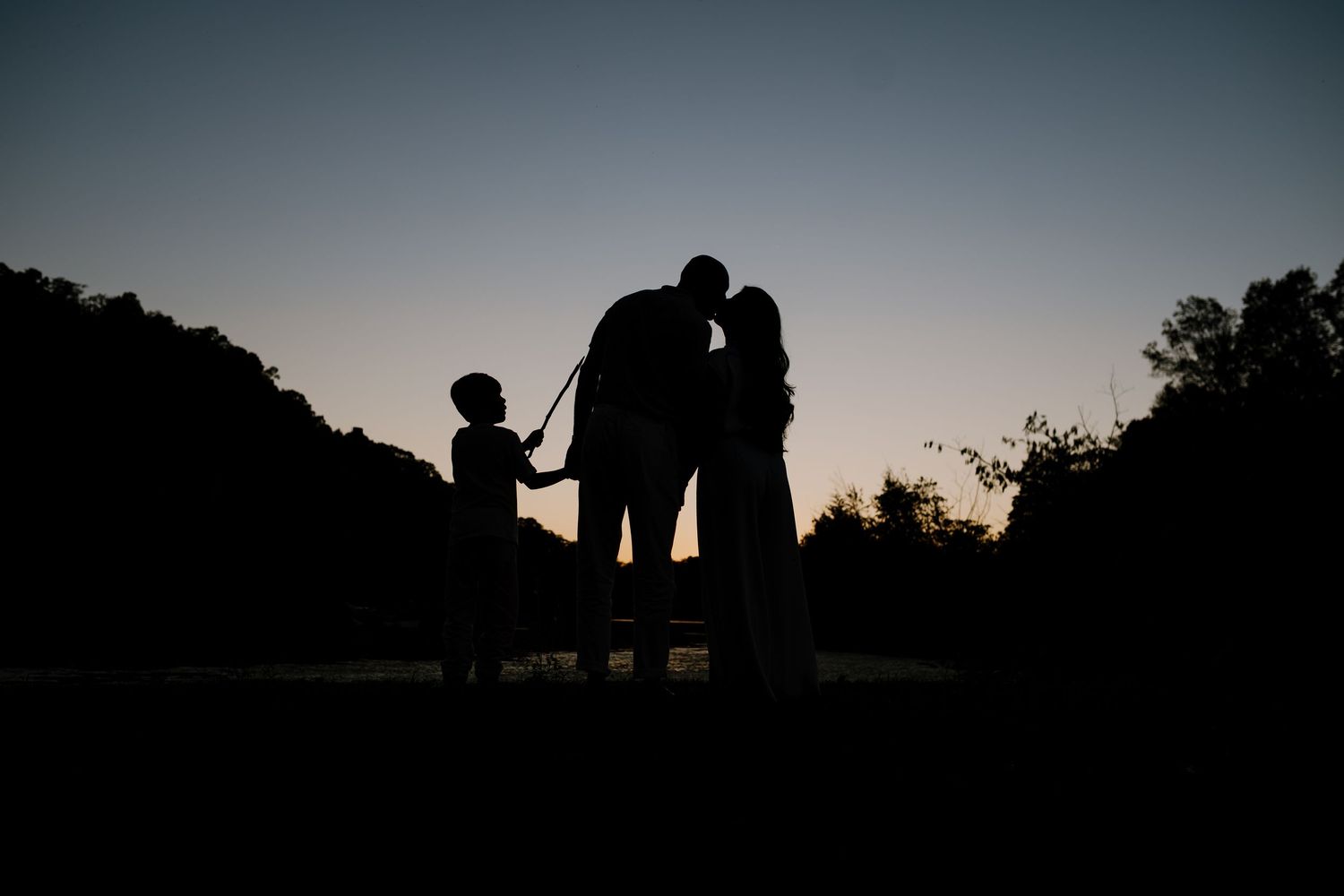 Silhouette of a family after sunset, parents are kissing and son points with a stick