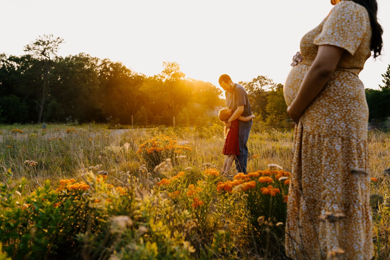 A dad hugs his daughter in a golden field of flowers with mom standing nearby watching, holding her pregnant belly
