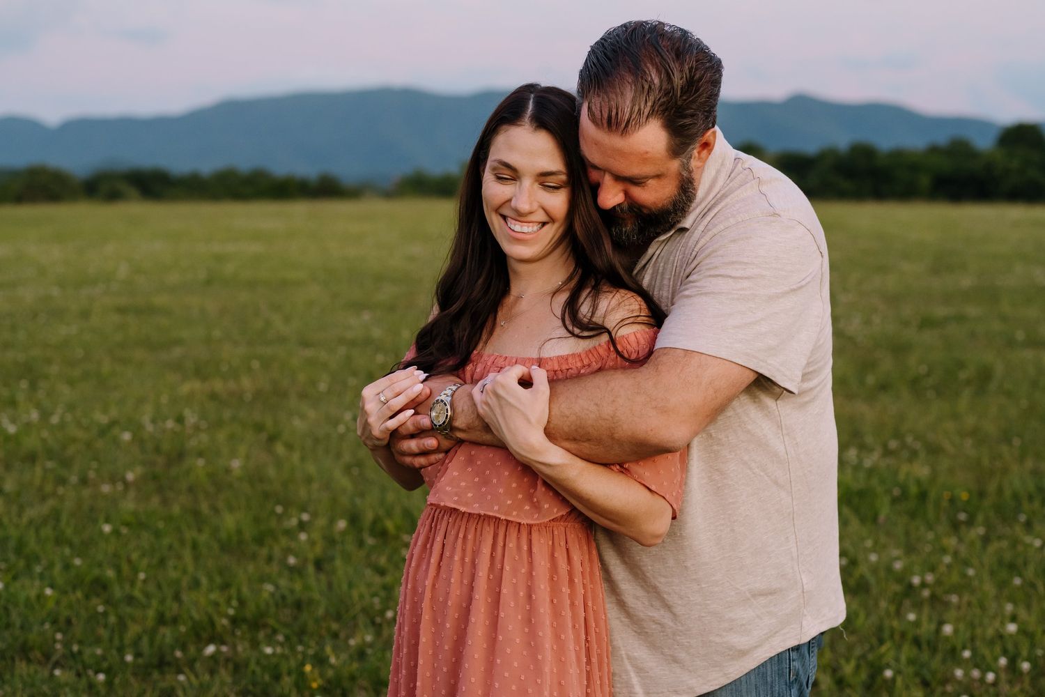 Couple embracing and smiling in a green field with mountain backdrop at sunset.