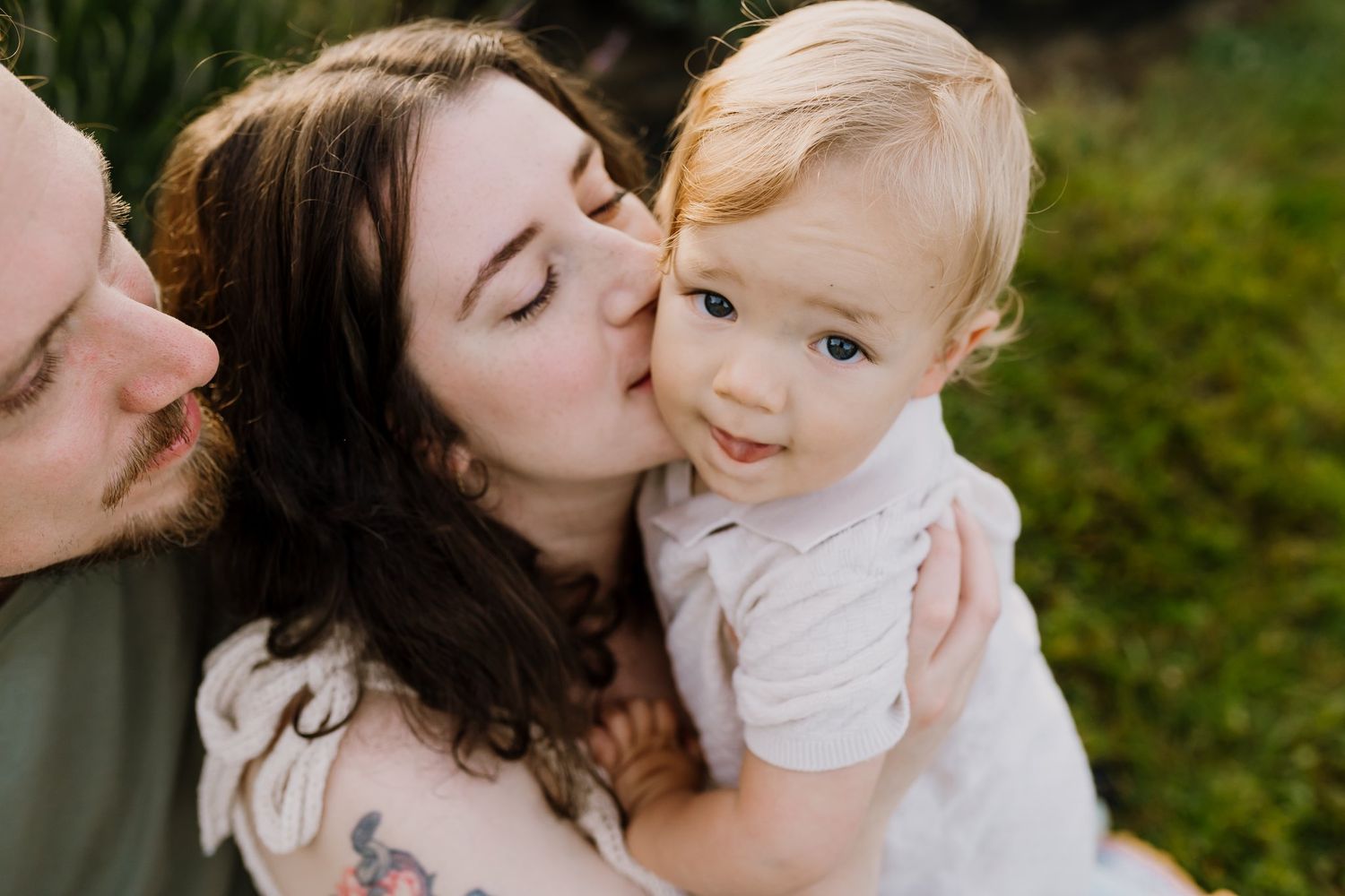 A mother tenderly kisses her smiling blonde toddler outdoors in a lush green garden setting.