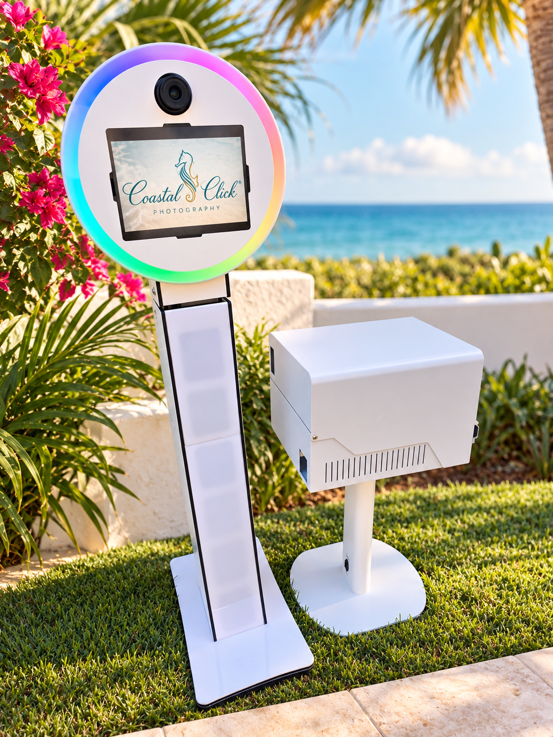 Coastal Click Photography photo booth with RGB ring light and printer stand on grass with ocean view backdrop.
