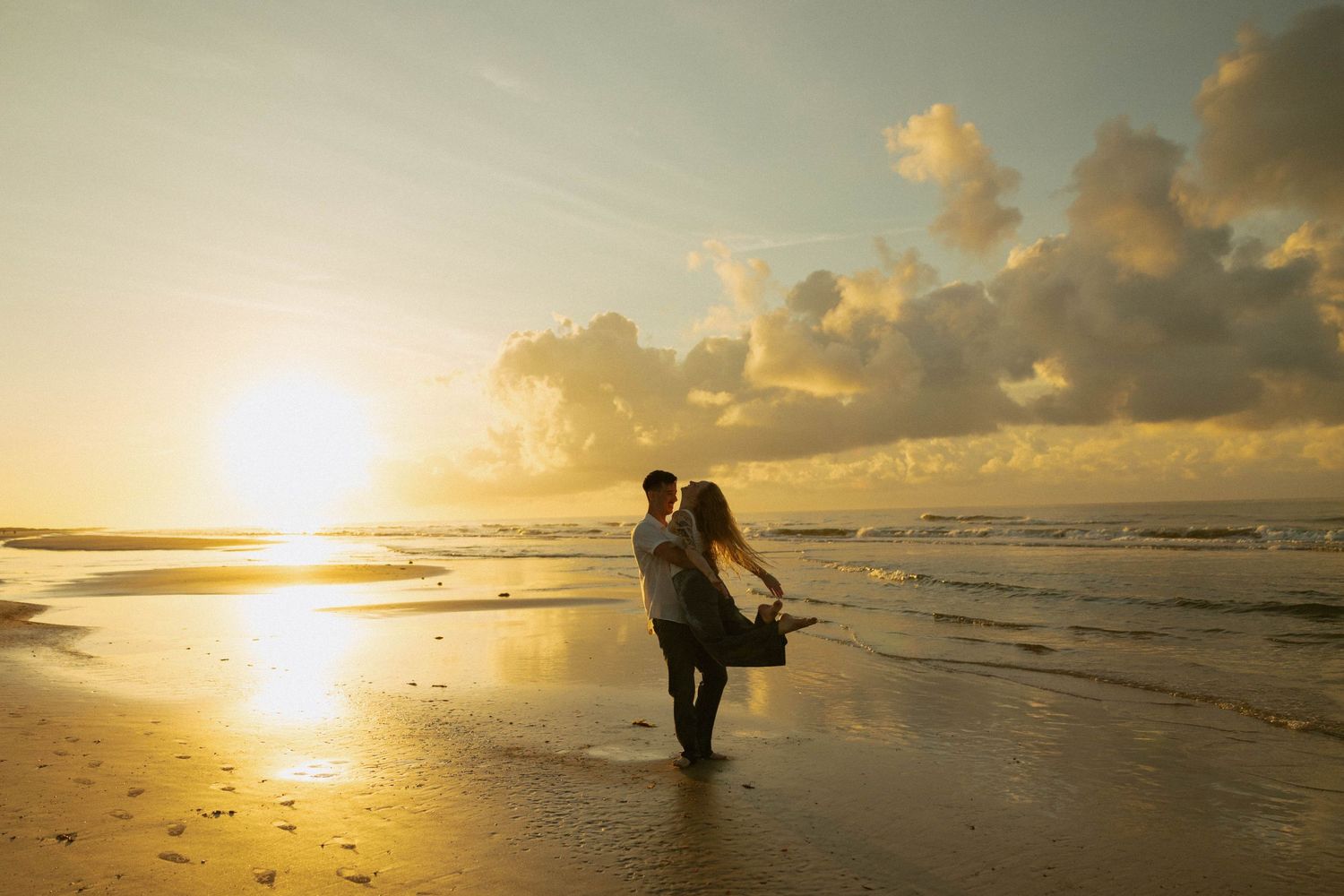 Couple embracing on a golden sandy beach at sunset, with waves and dramatic clouds in the background.