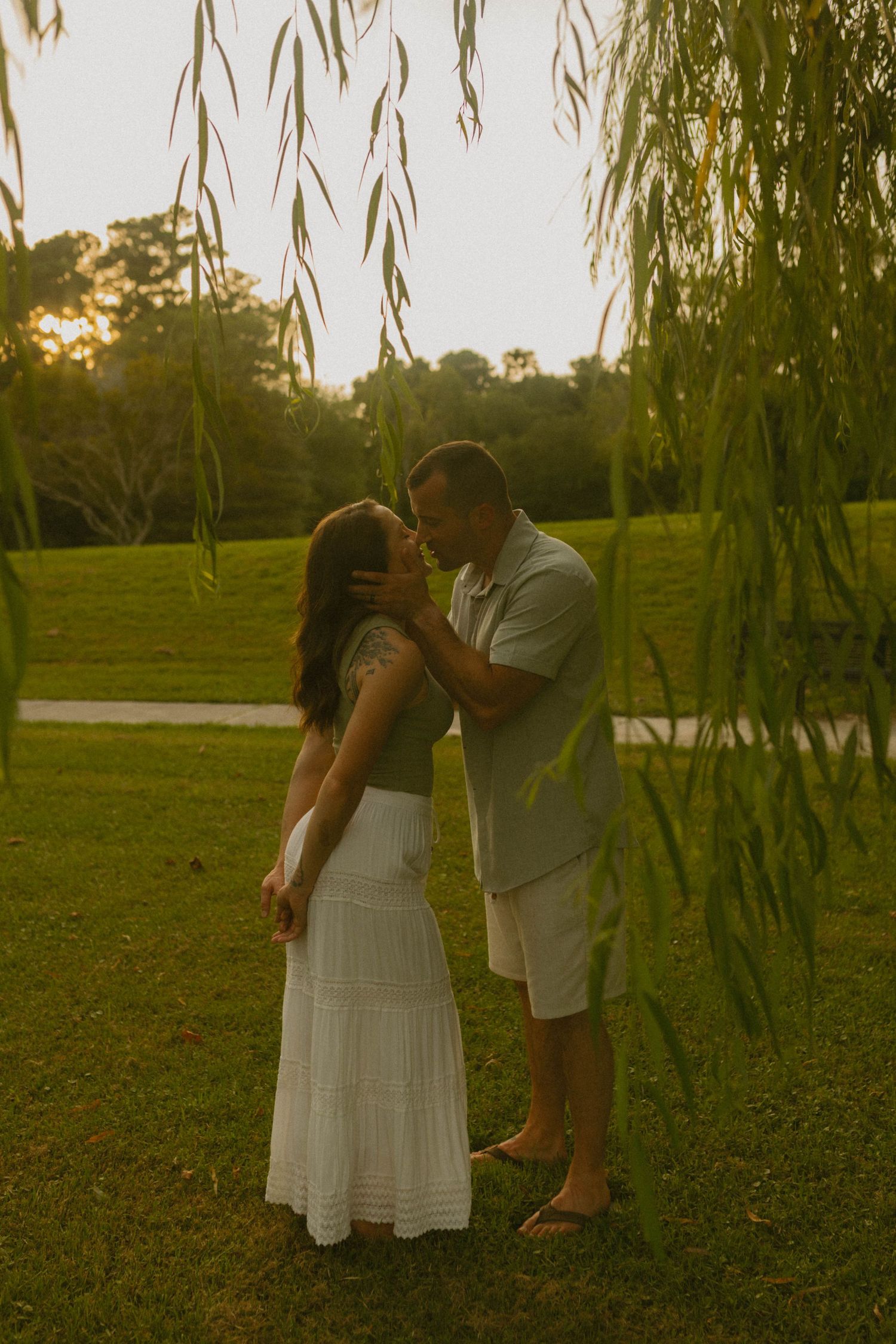 Couples photographer capturing a romantic moment at sunset under drooping willow branches in a lush green park in Jacksonville, NC