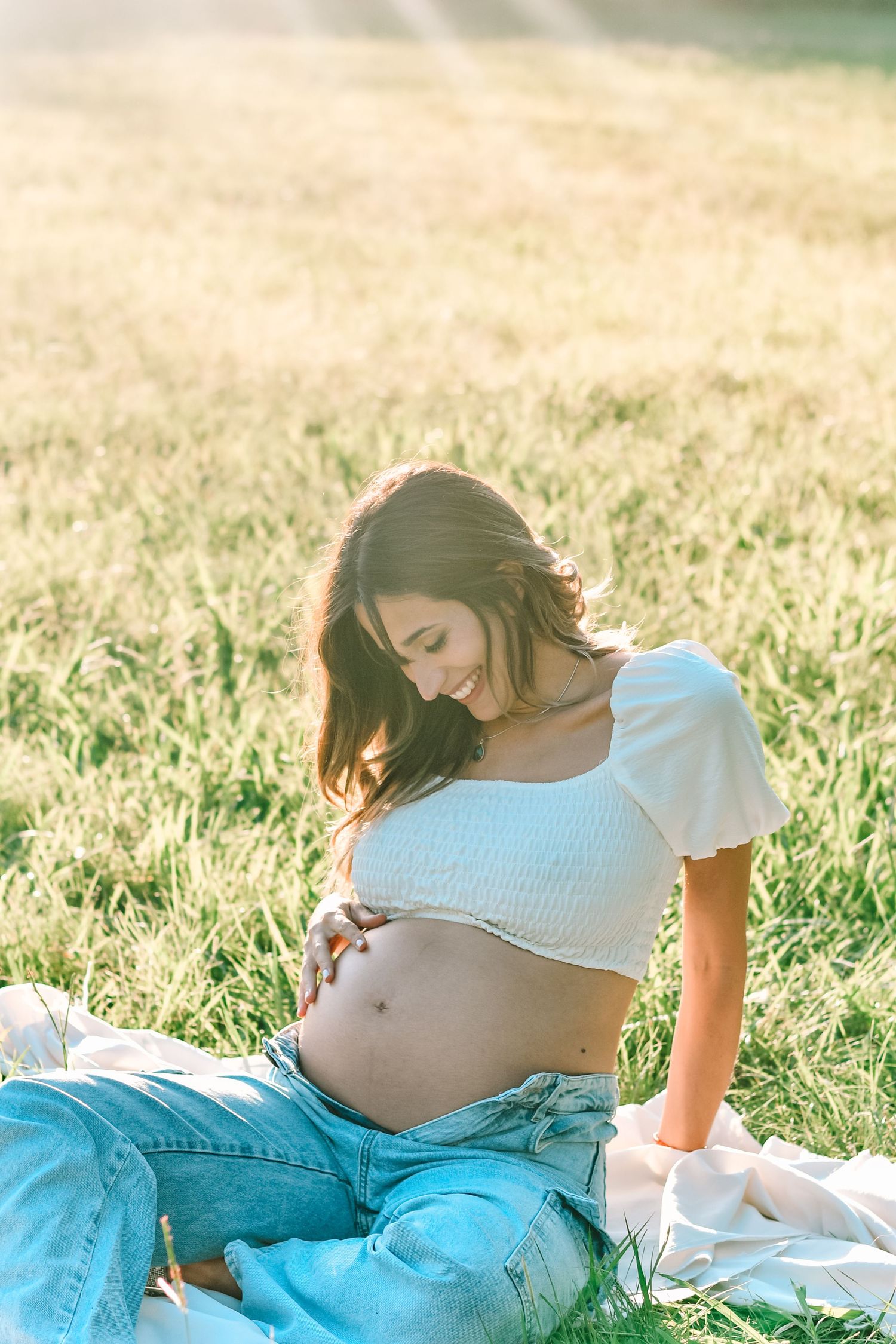 Maternity photo in sunlit field with white crop top and denim during golden hour.