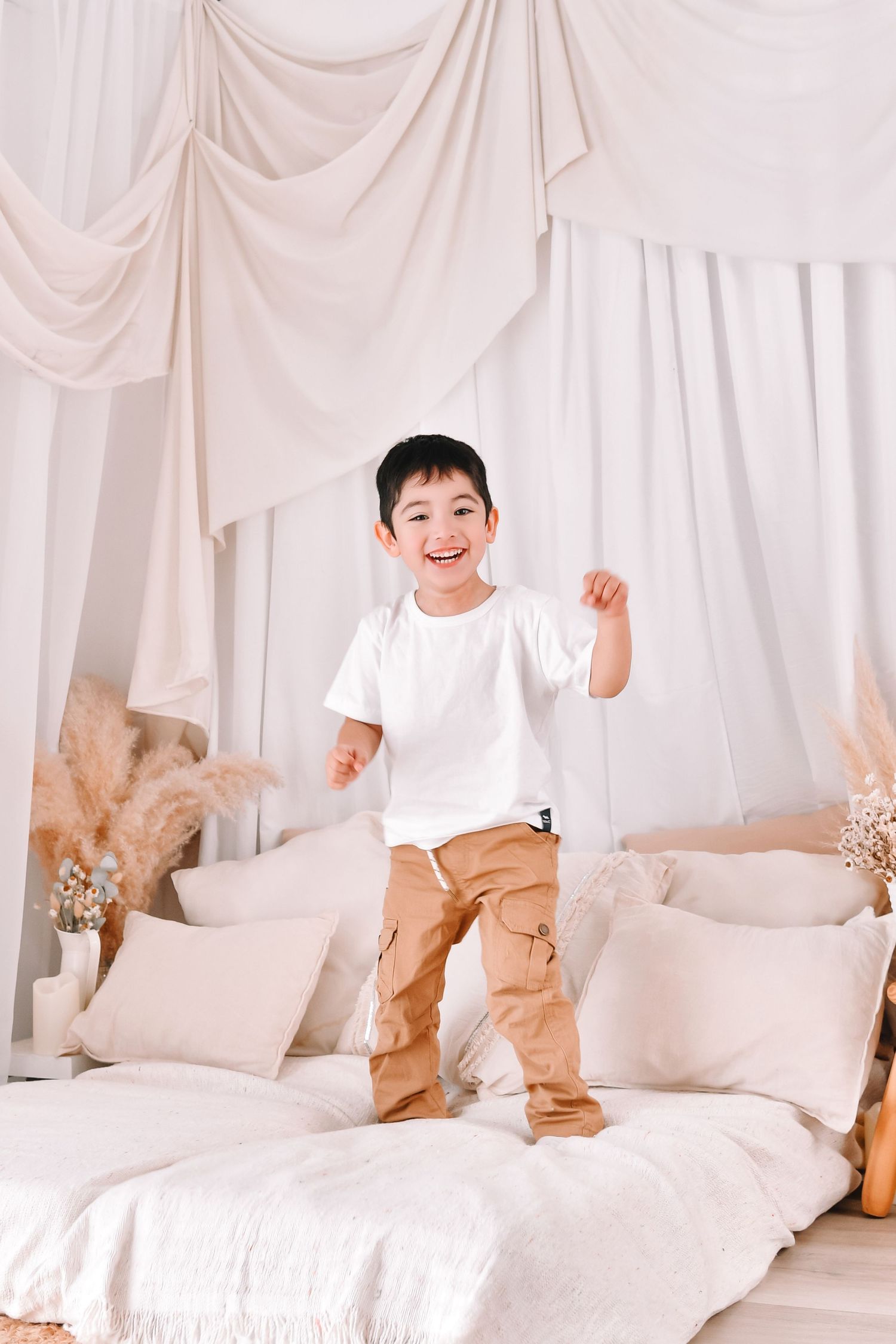 Young boy in white shirt and tan pants jumping playfully on a white bed with decorative drapes and pillows.