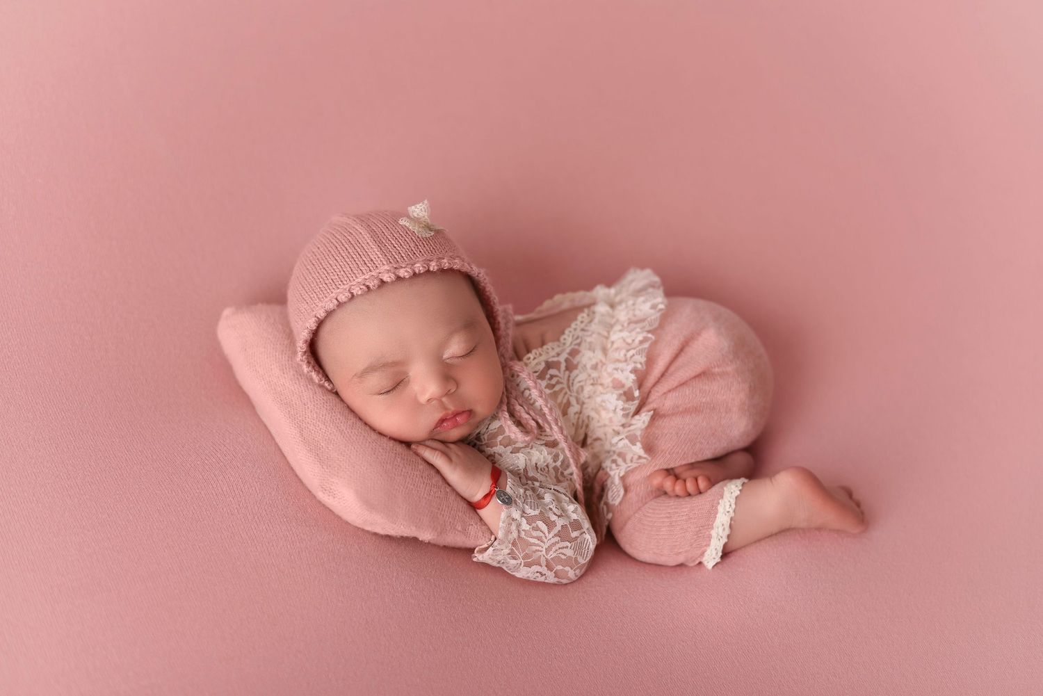 Sleeping newborn in pink lace outfit curled up on soft pink pillow.