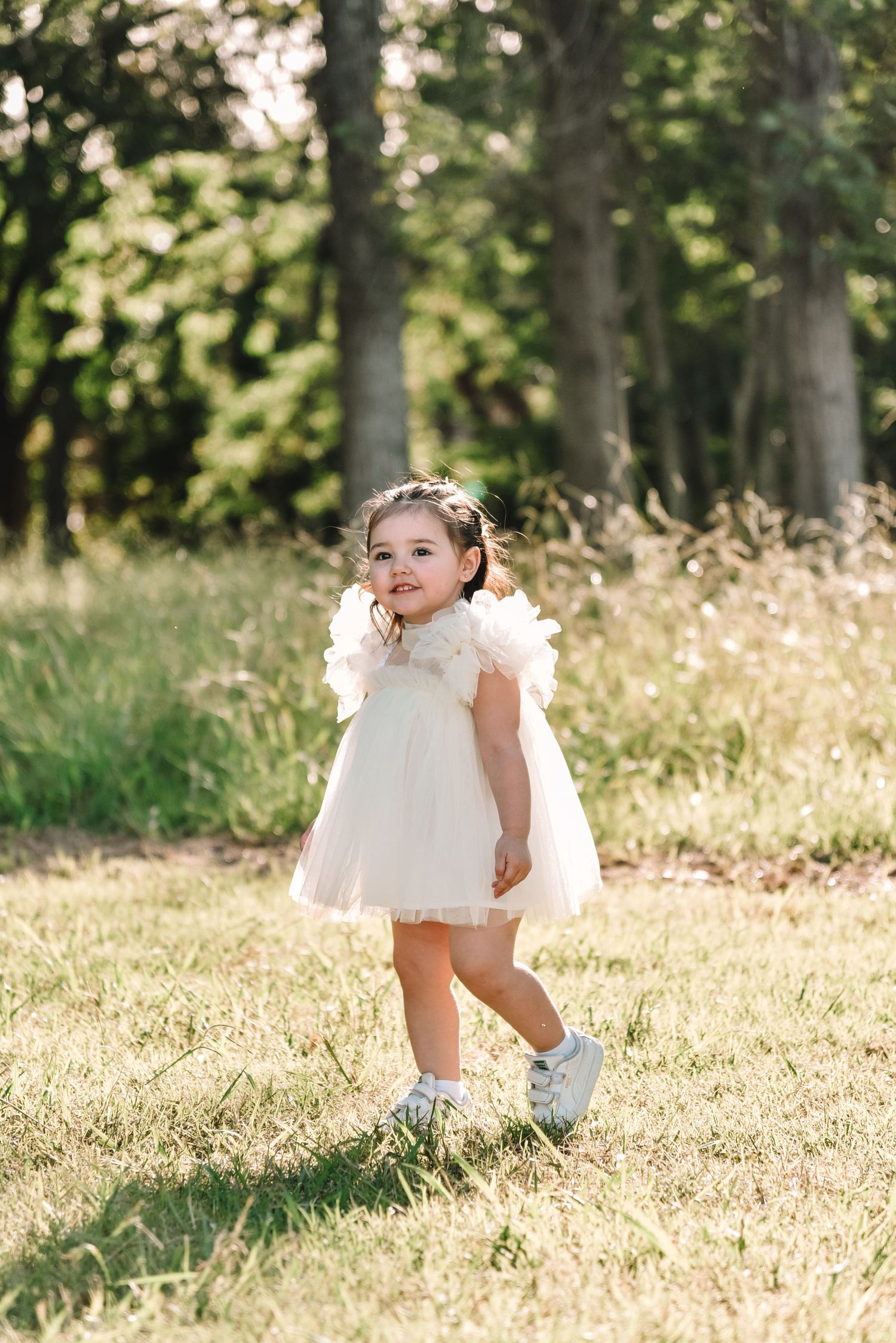 Sunlit outdoor portrait in a grassy field with white dress and feather details.