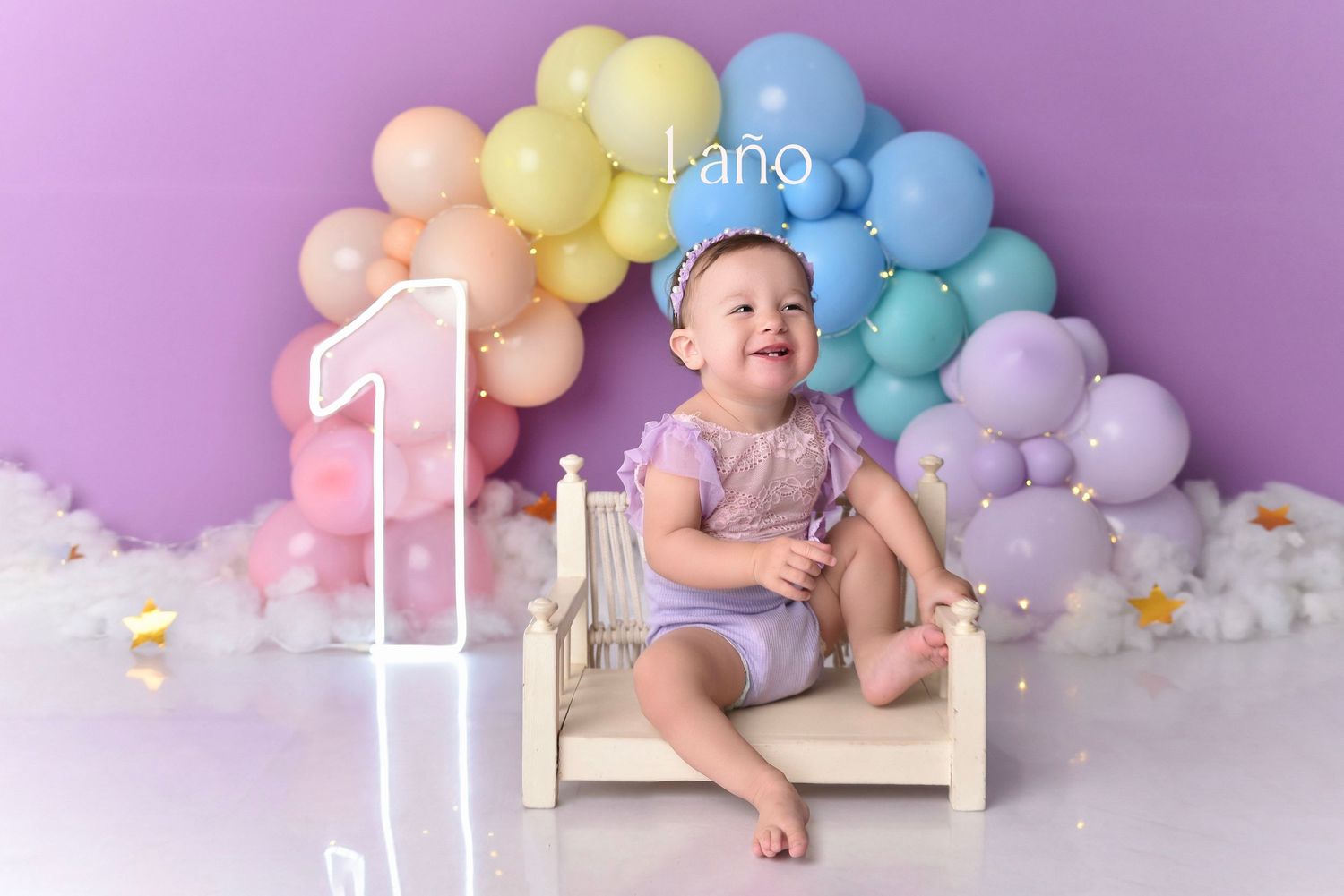 First birthday celebration setup with pastel rainbow balloon arch and white number one against purple backdrop.