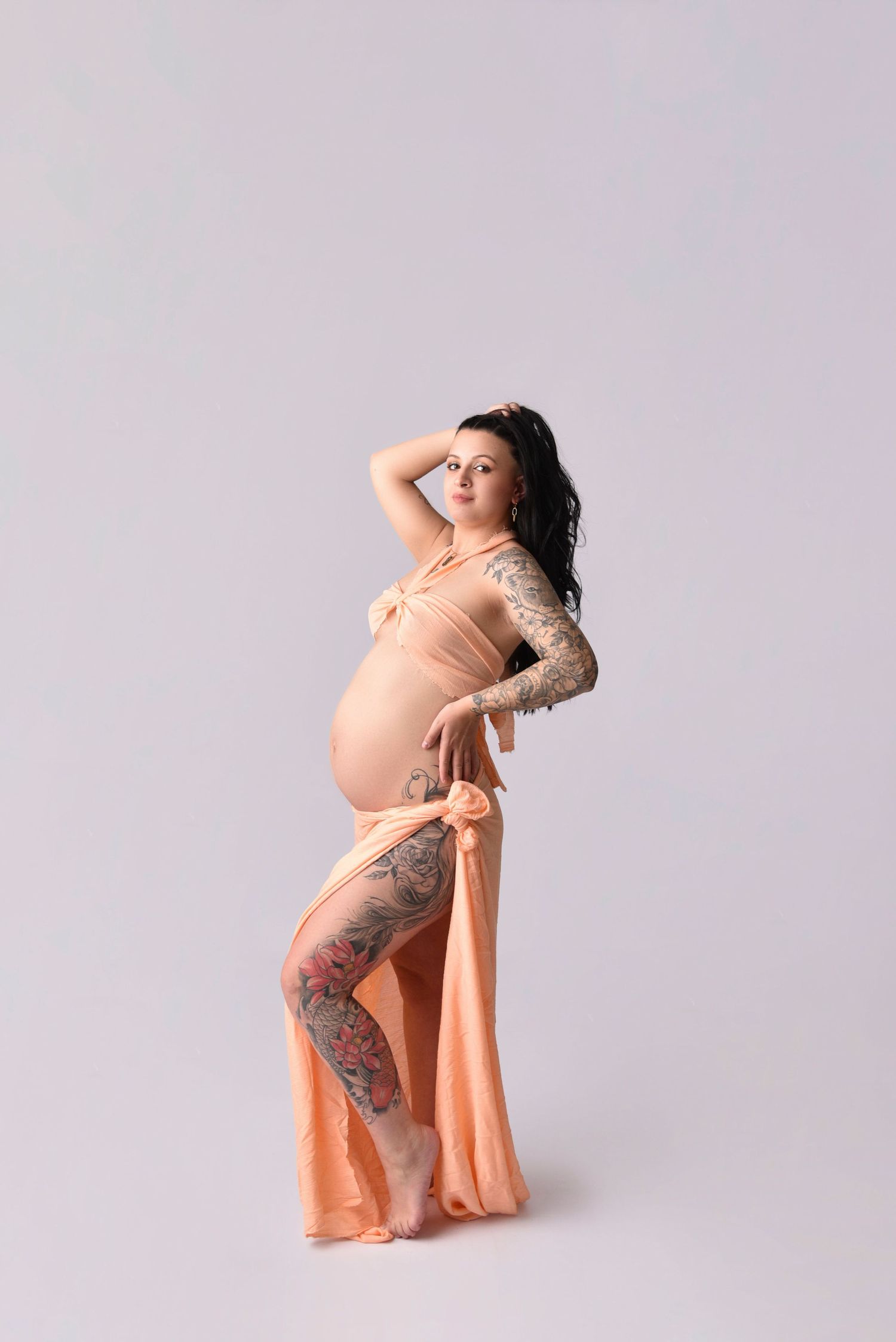 Artistic maternity portrait with flowing peach fabric showcasing intricate leg tattoo against light background.