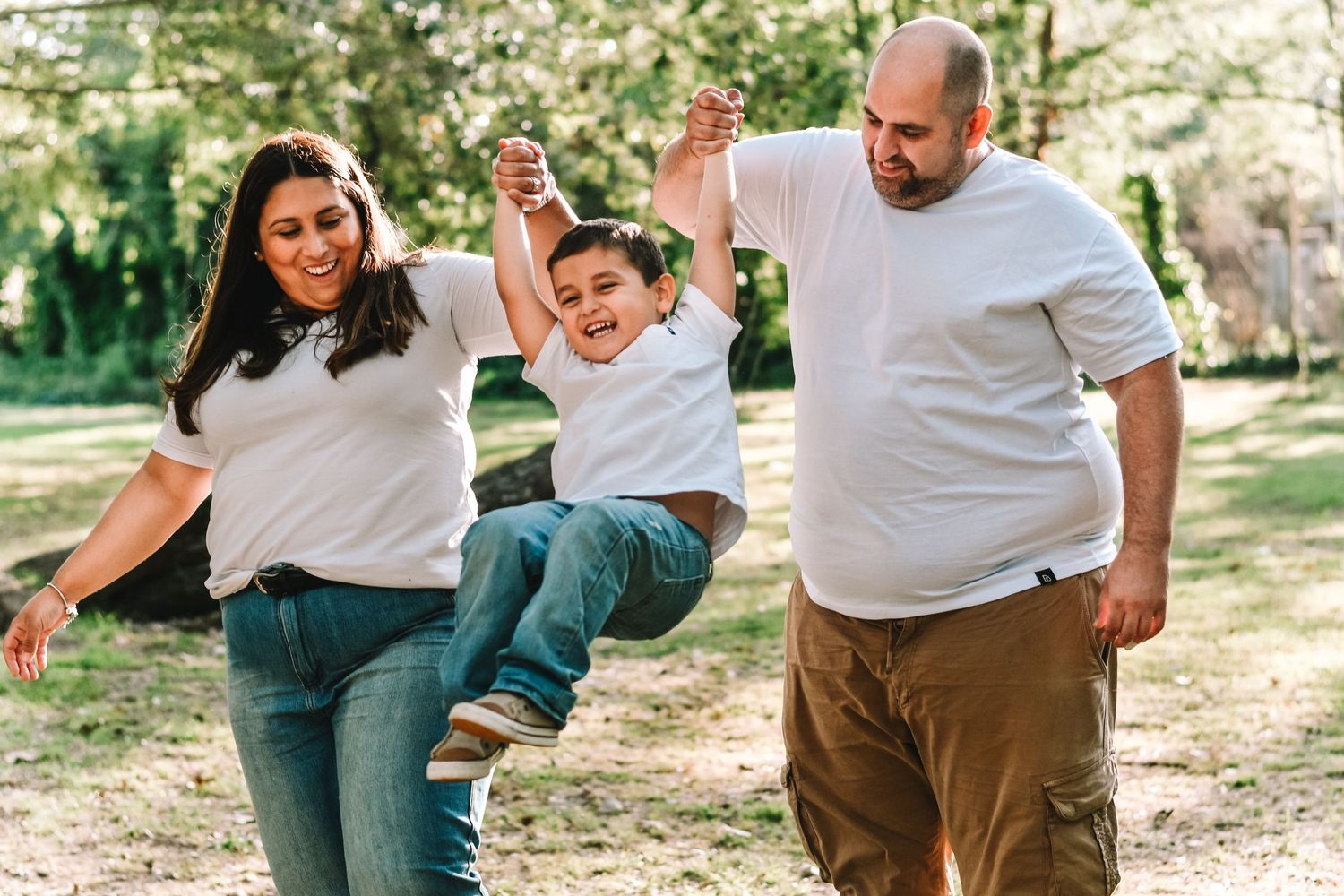Family plays with child in park, lifting little one while all wearing casual white tops.