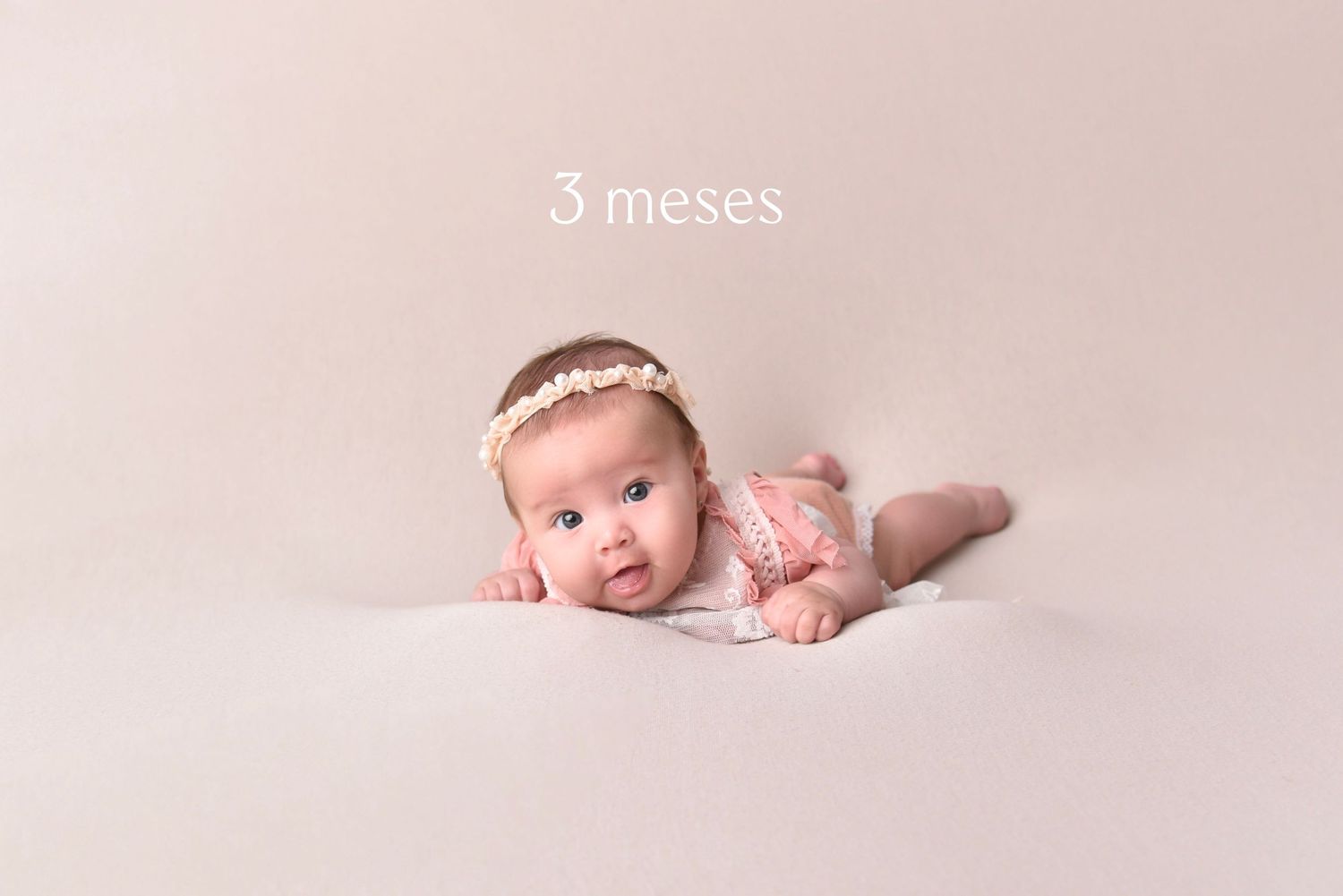 A tiny baby wearing a pink lace outfit and headband lies on their tummy on a beige backdrop during a 3-month photoshoot.