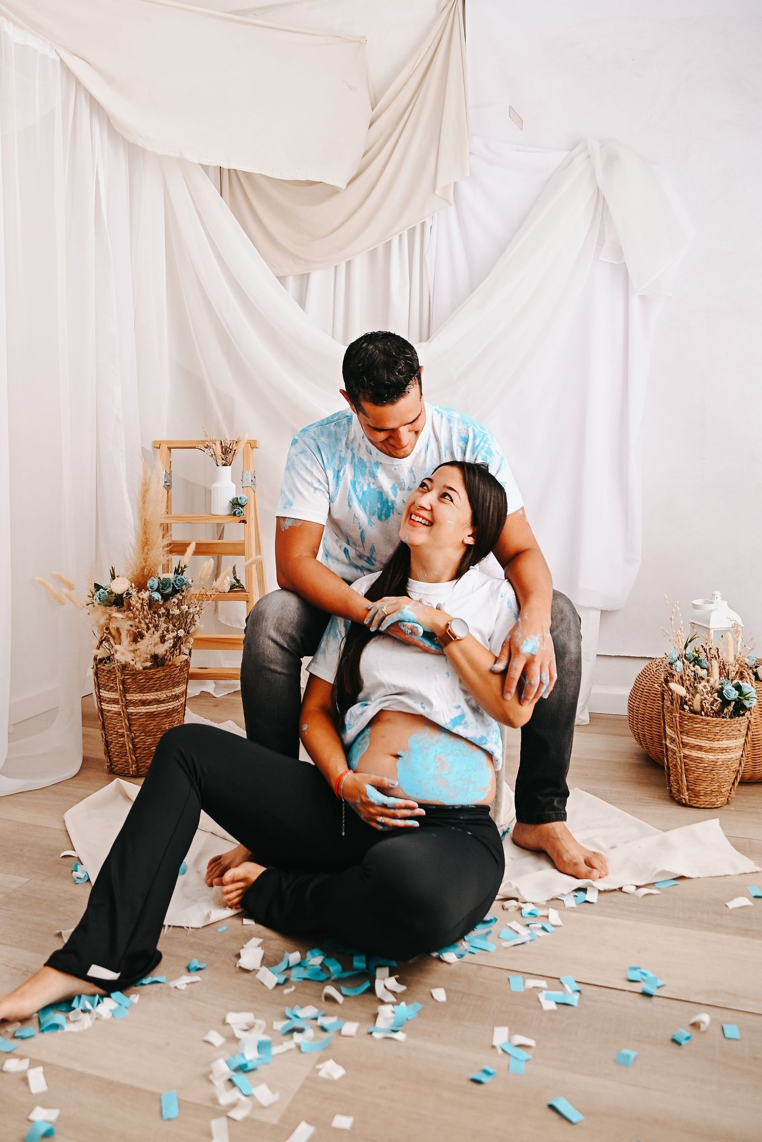 Couple celebrating gender reveal with blue confetti, pregnant woman sitting on floor with partner behind her in decorated studio.