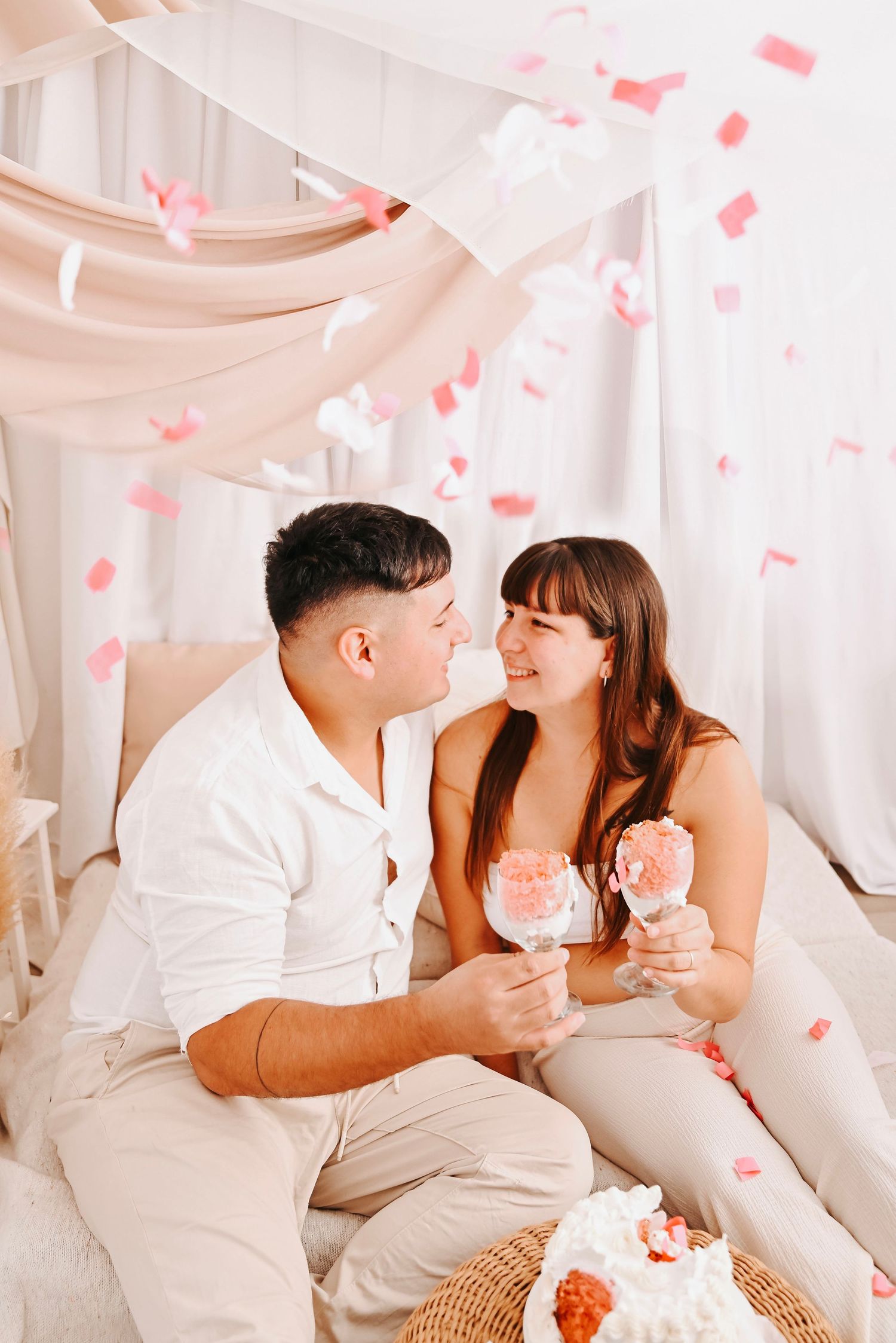 Couple sitting together smiling, holding pink drinks with confetti falling around them in a romantic pink decorated setting.