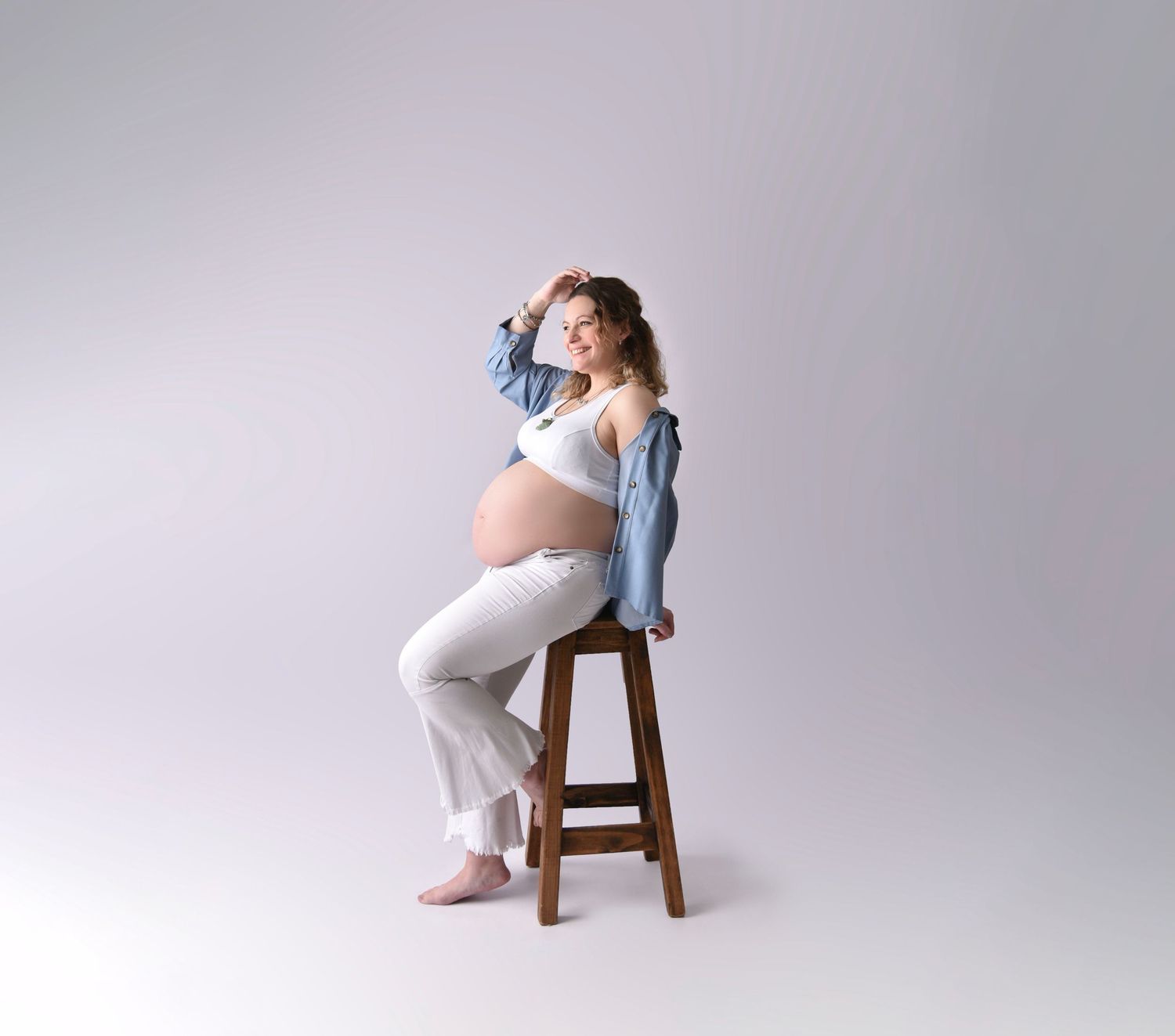 Maternity portrait of an expectant mother in white loungewear and denim jacket sitting on wooden stool against gray backdrop.