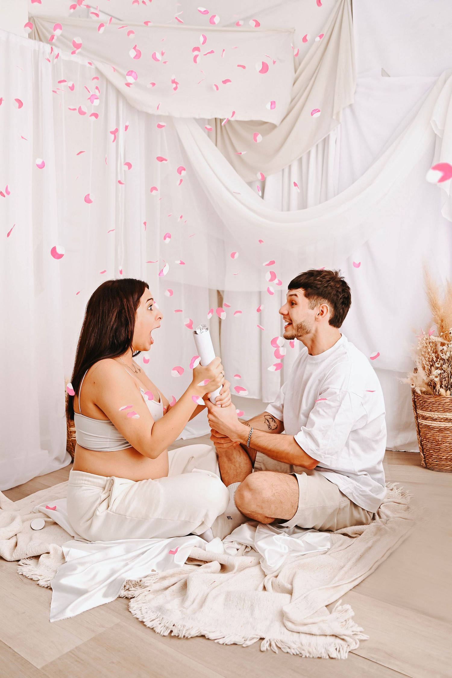Couple sitting on floor with pink confetti falling, celebrating a gender reveal with a confetti popper.