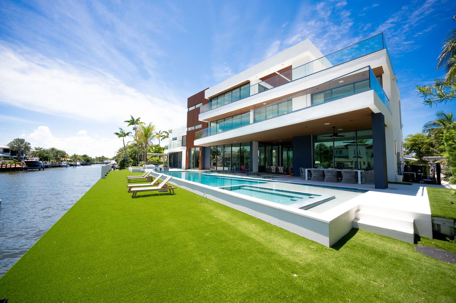 Modern luxury waterfront home with infinity pool, lush green lawn, and glass facade overlooking a serene canal on a sunny day.