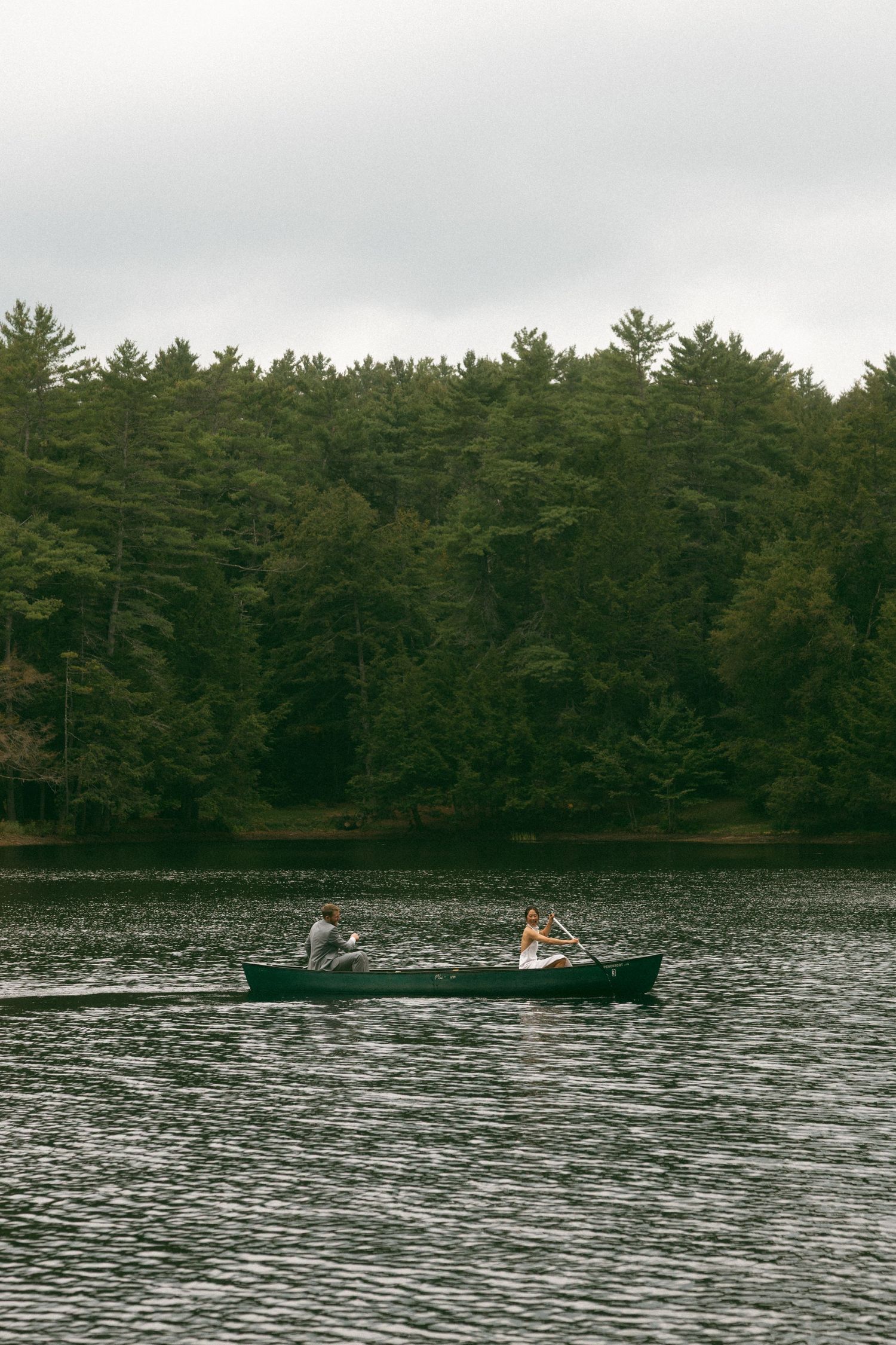 People paddling a canoe across a serene lake surrounded by dense pine forest on an overcast day.