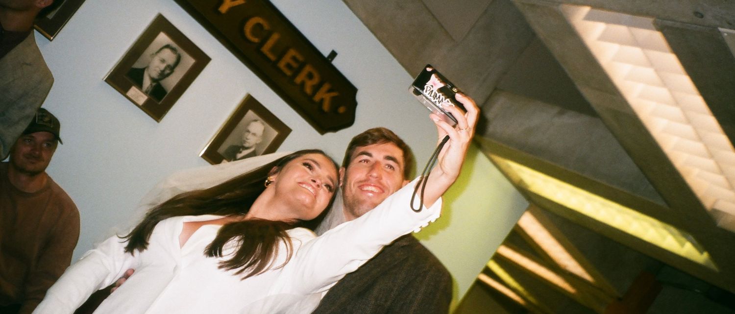 Two people taking a selfie with a digital camera indoors near a Clerk sign.