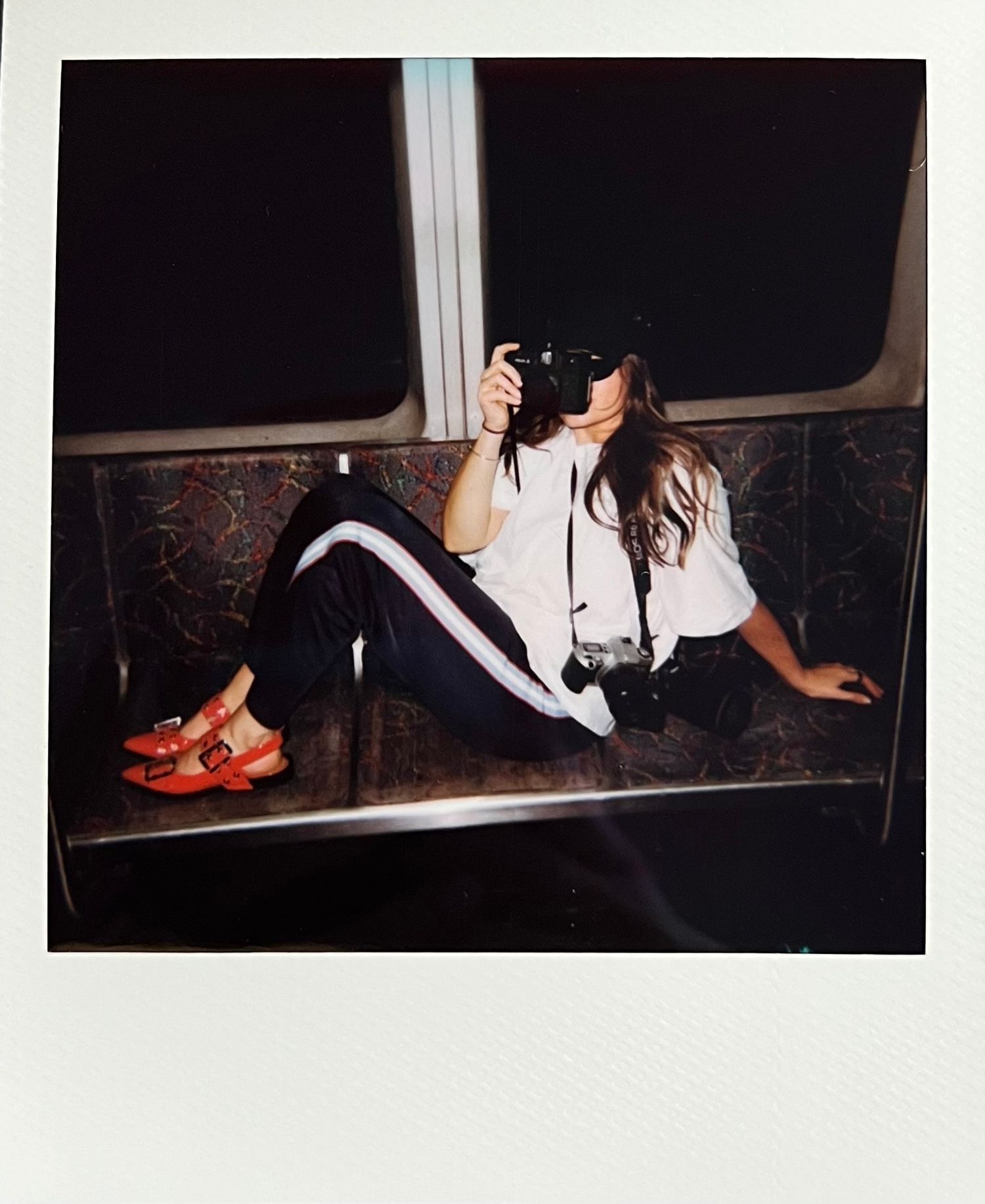 Woman sitting on subway bench taking photo with camera, wearing white shirt, striped pants, and red shoes.