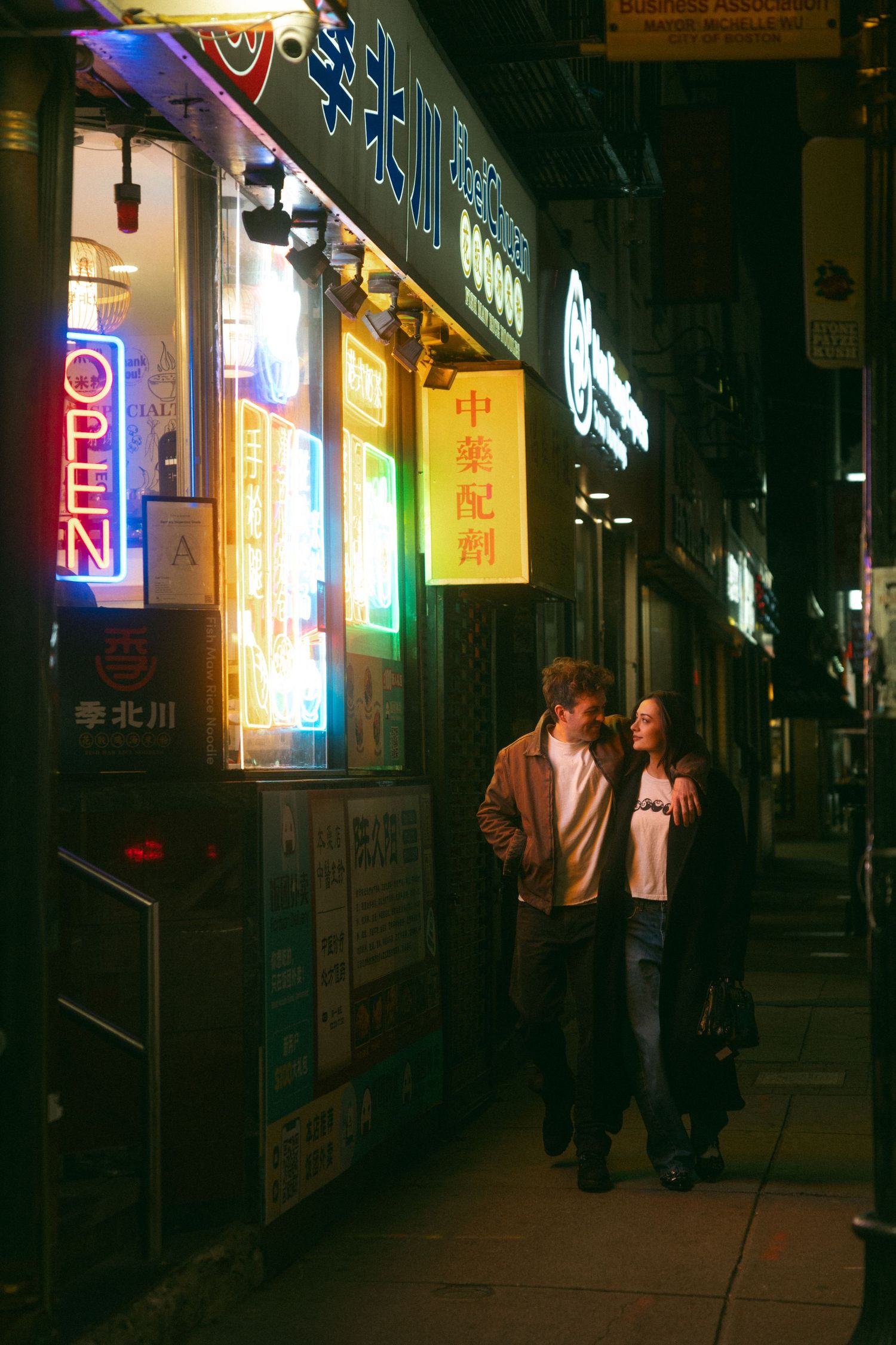 Couple walking past neon-lit Chinatown storefronts at night, colorful Chinese signs glowing on urban sidewalk.