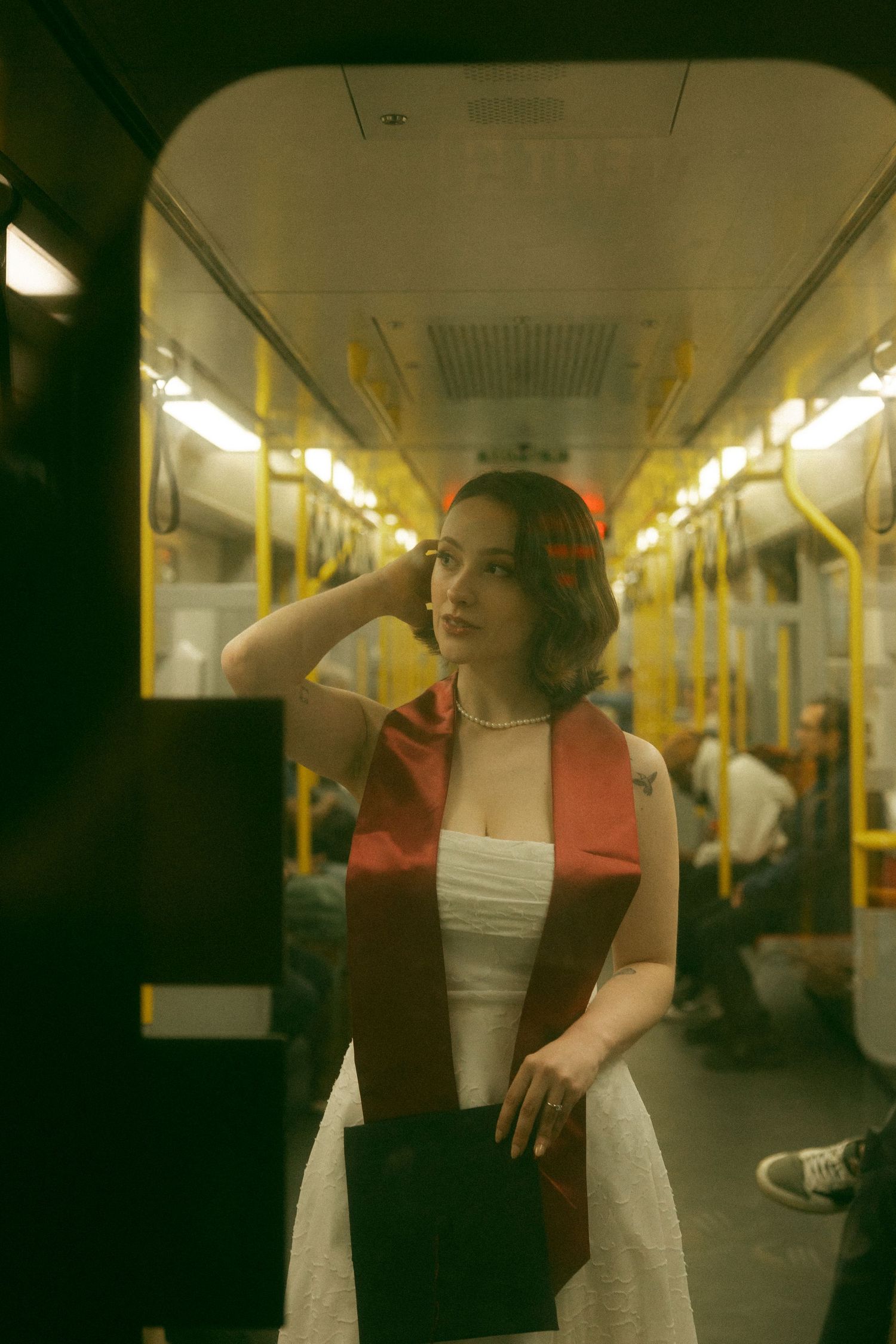 A young woman in a white dress and red sash stands in a subway car, holding a book while passengers sit behind her.
