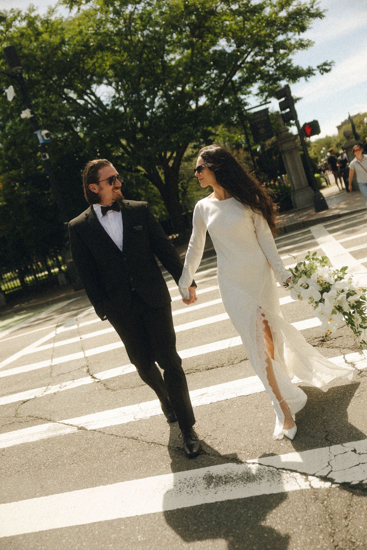 Bride and groom in elegant wedding attire holding hands while crossing a sunny street crosswalk.
