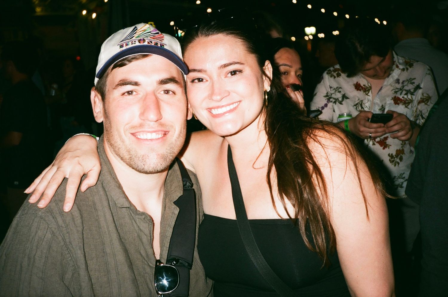 Two smiling friends pose together at a dimly lit social event, man in cap and olive shirt, woman in black top.