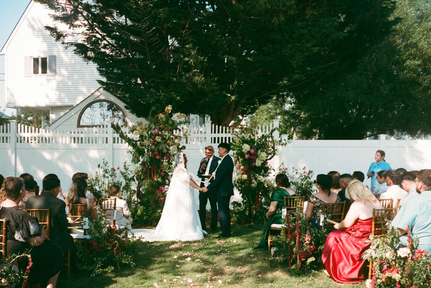 An outdoor garden wedding ceremony with guests seated on chairs surrounded by greenery and a white picket fence.