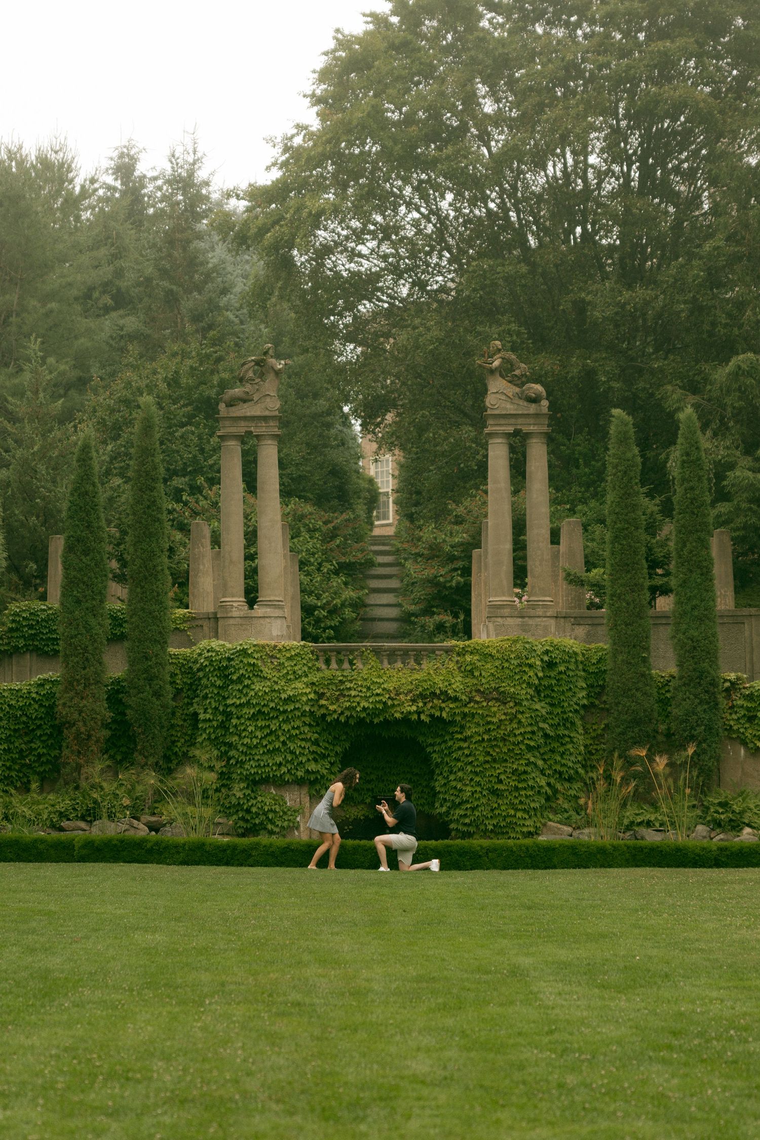 A couple shares a proposal moment on a lush green lawn surrounded by ivy walls and stone columns in a formal garden.