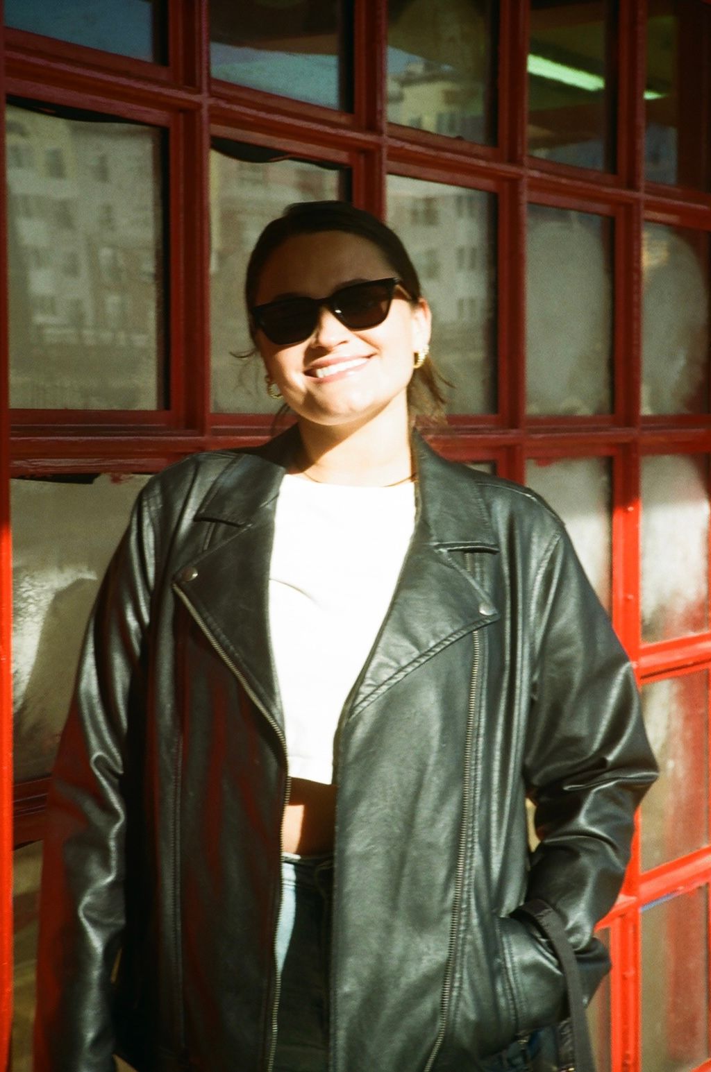 Smiling woman in black leather jacket and sunglasses posing in front of red-framed windows outdoors.