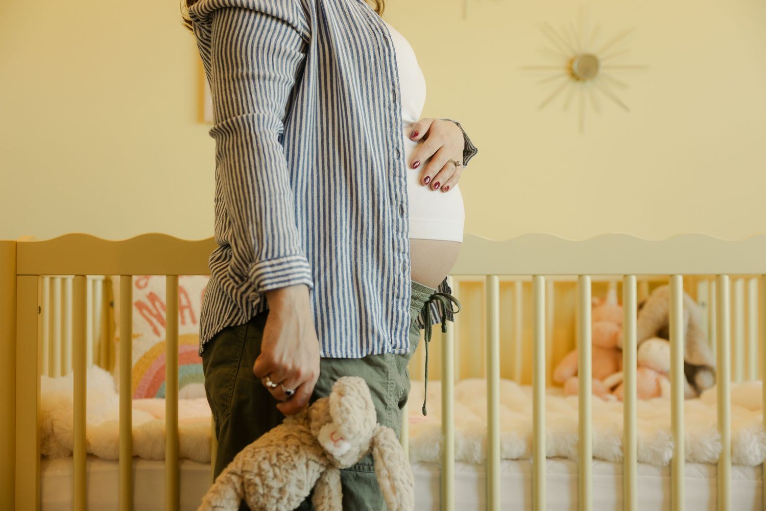 Pregnant woman in striped shirt holding teddy bear stands beside a wooden nursery crib in a warm yellow room.