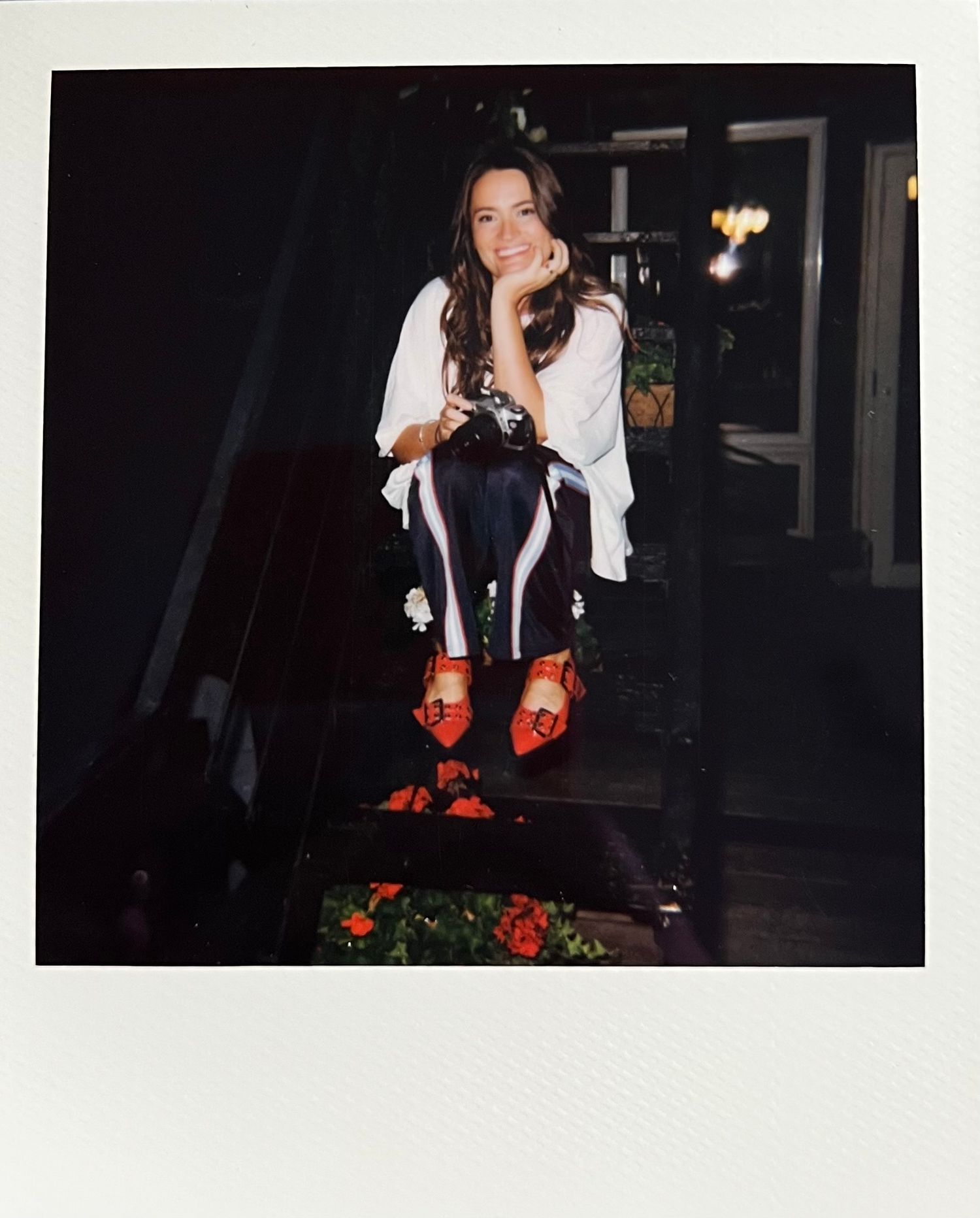Woman in white blouse and dark floral pants with red shoes sitting on steps in a Polaroid-style photo.