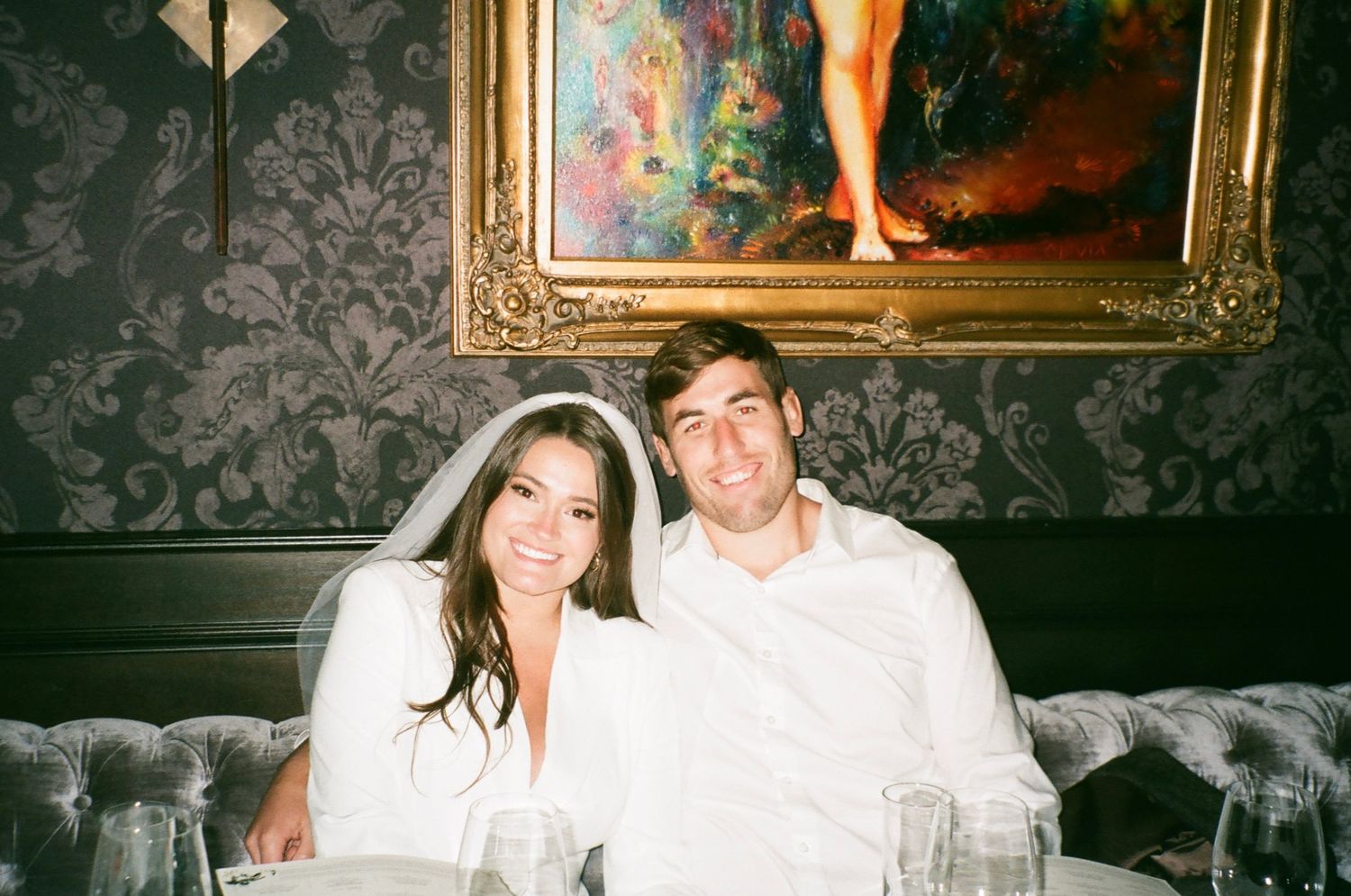 Bride and groom smiling together in elegant venue booth beneath ornate gold-framed painting on dark wallpaper.