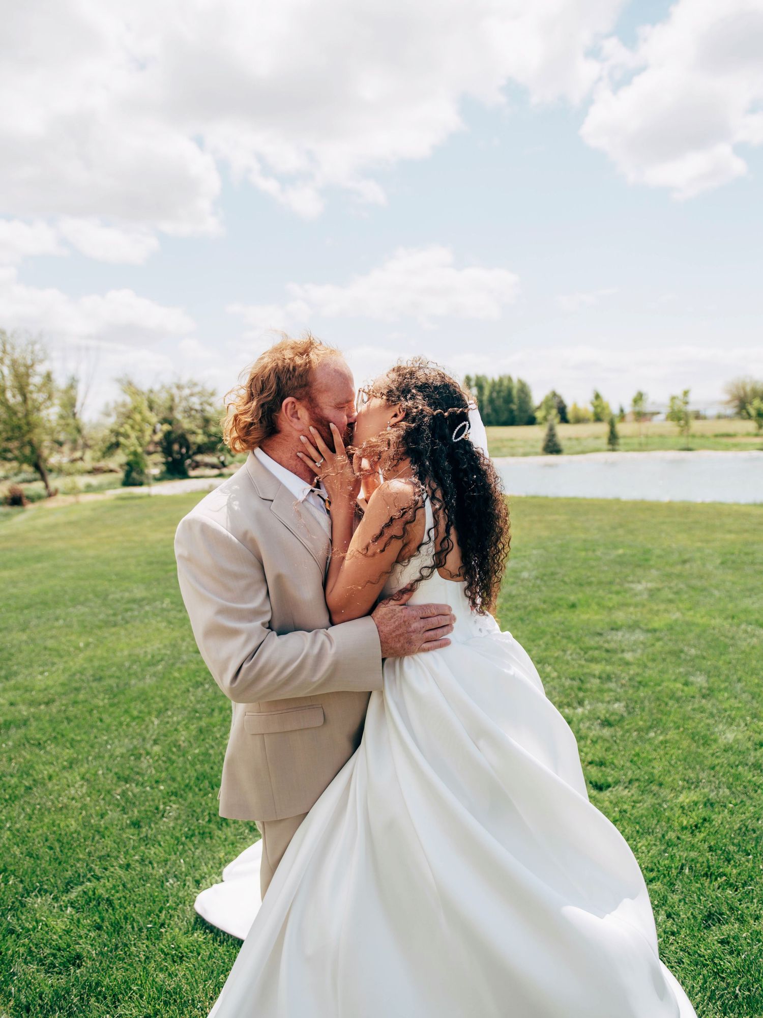 A romantic outdoor wedding photo of a couple embracing on a grassy field with a lake in the background.