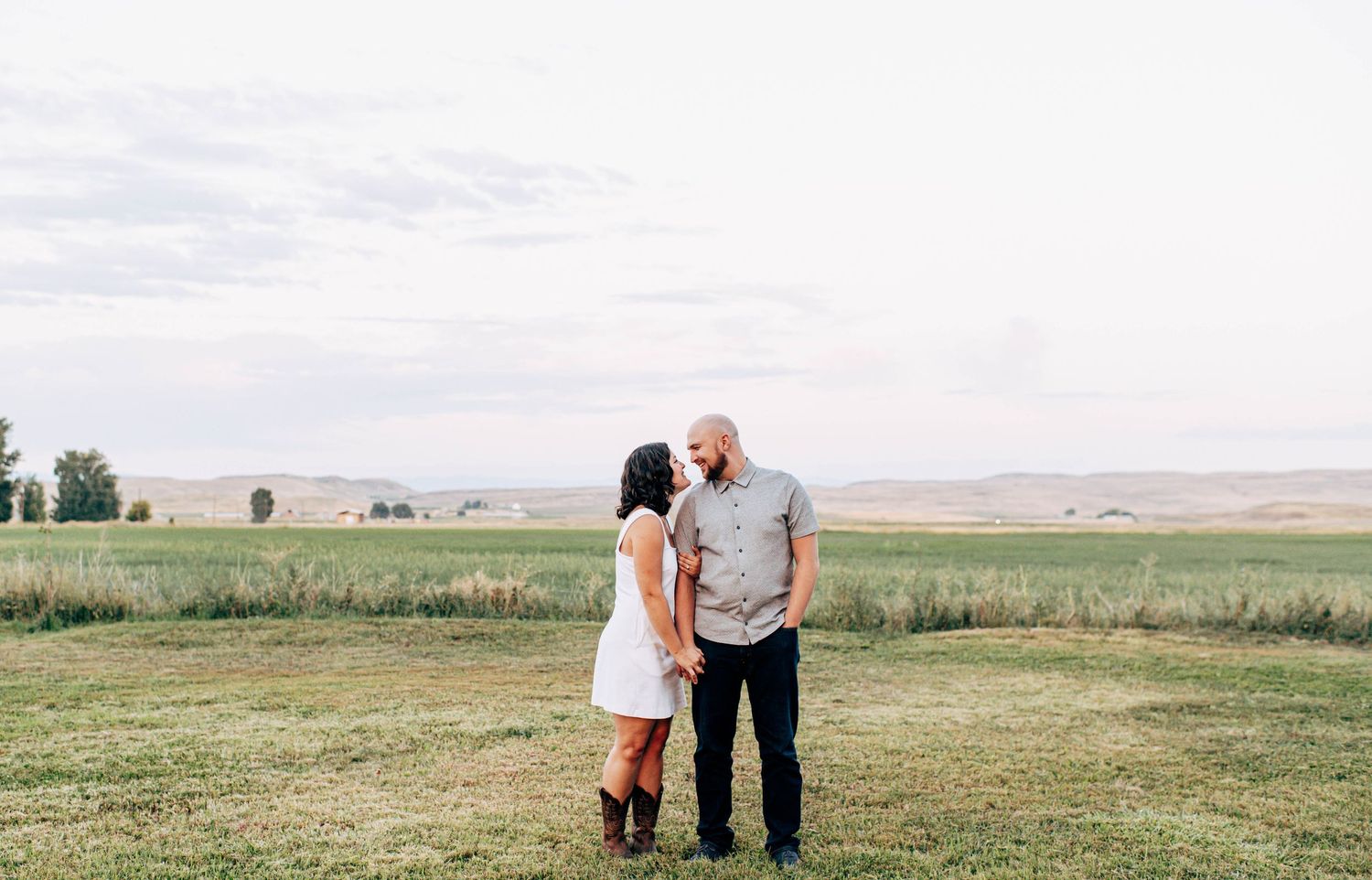 Couple holding hands and gazing at each other in a lush green field with rolling hills and open sky.