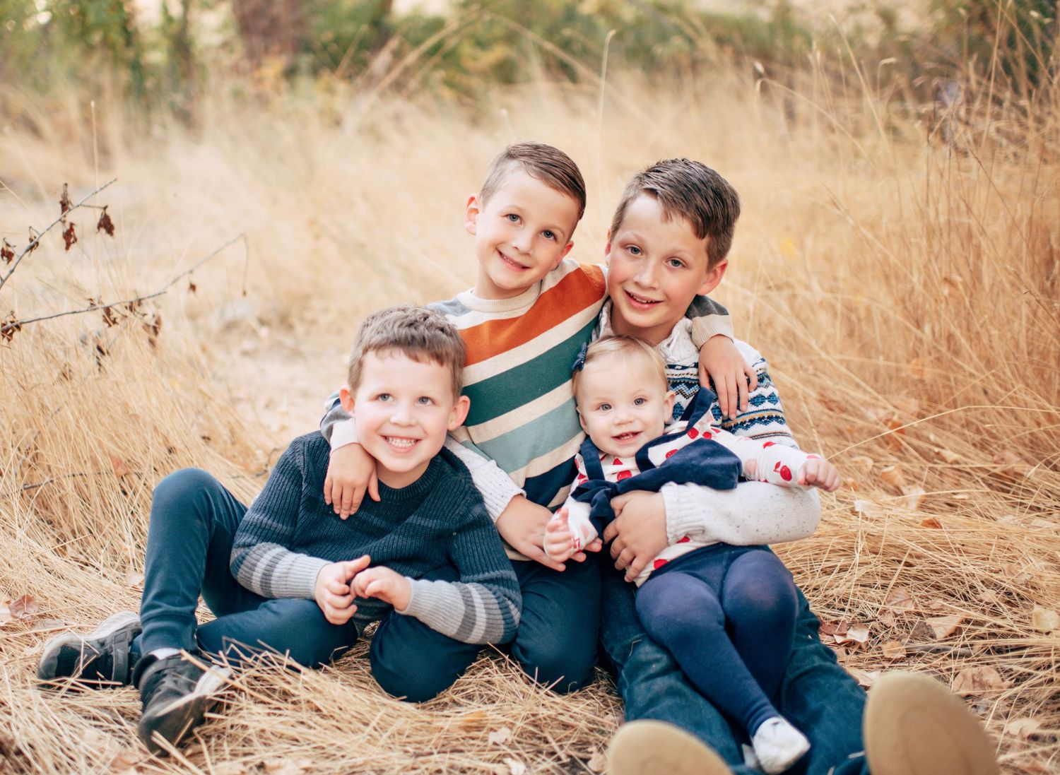 Four smiling children sitting together in a golden field, wearing colorful fall outfits, with dry grass in the background.
