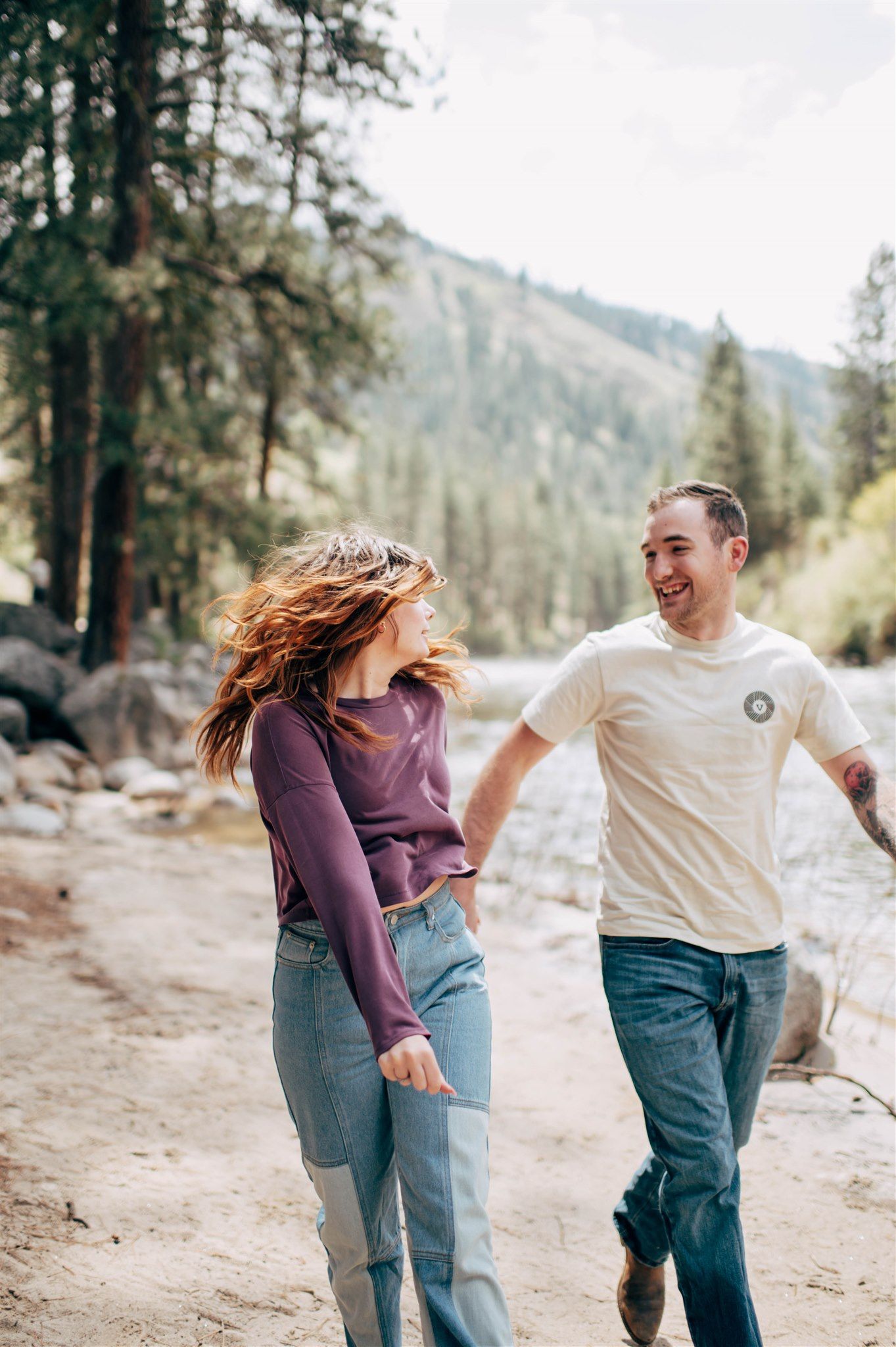 A joyful couple laughs and holds hands while walking along a scenic river trail surrounded by pine trees.