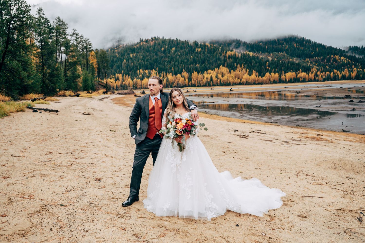 Bride and groom pose on sandy lakeside beach with autumn trees and misty mountains in background.