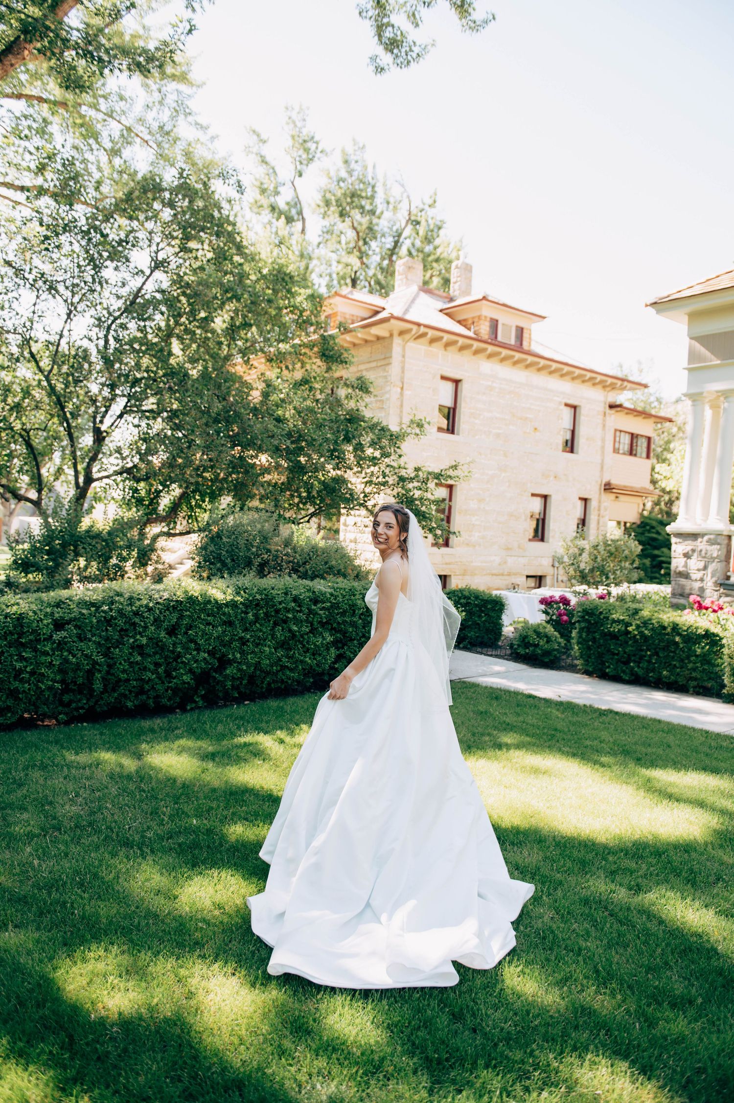A bride in a flowing white wedding gown twirls on green lawn outside an elegant stone mansion surrounded by lush hedges.