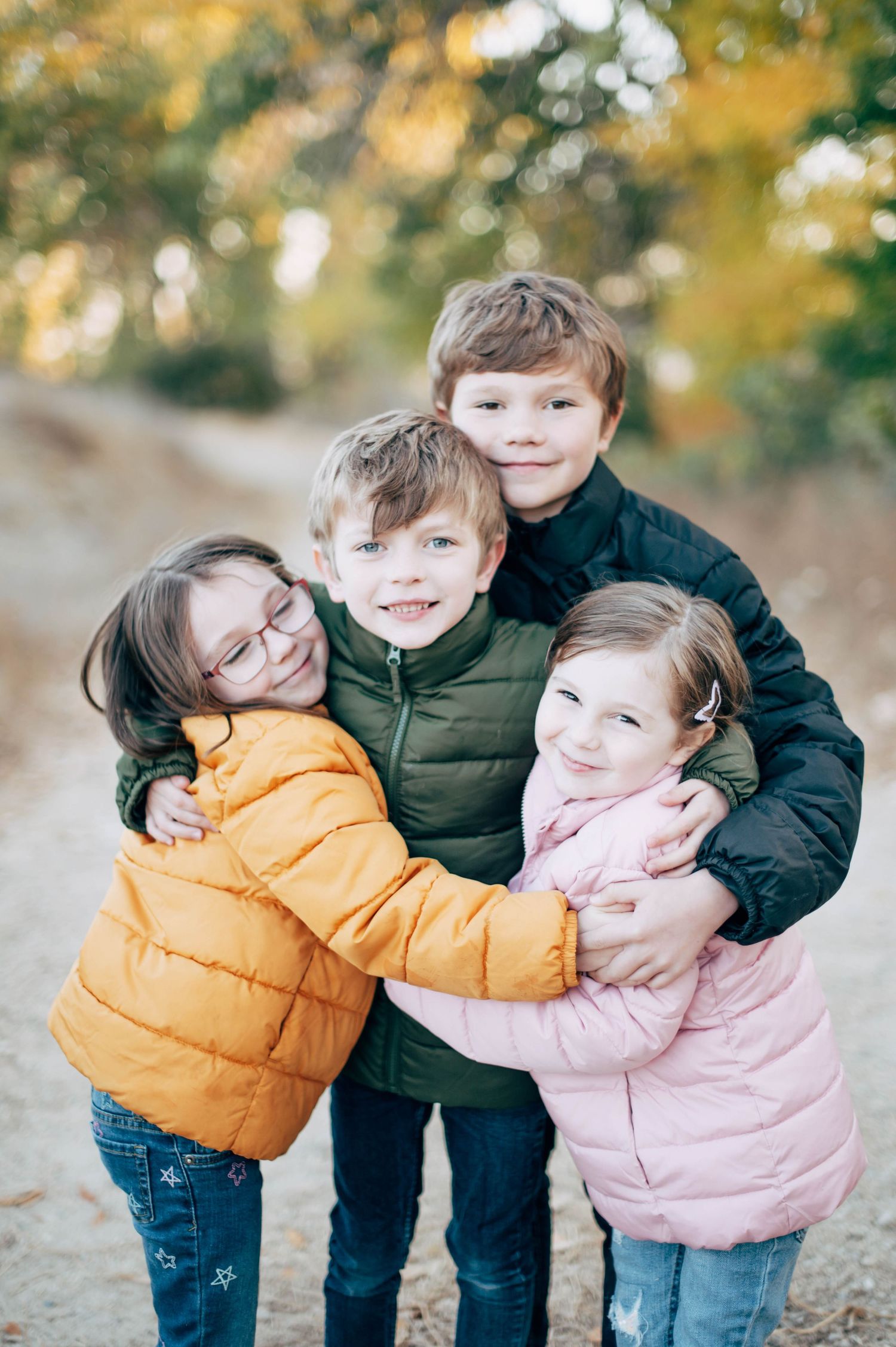 Four children in colorful winter jackets hugging outdoors on a dirt path surrounded by autumn foliage.