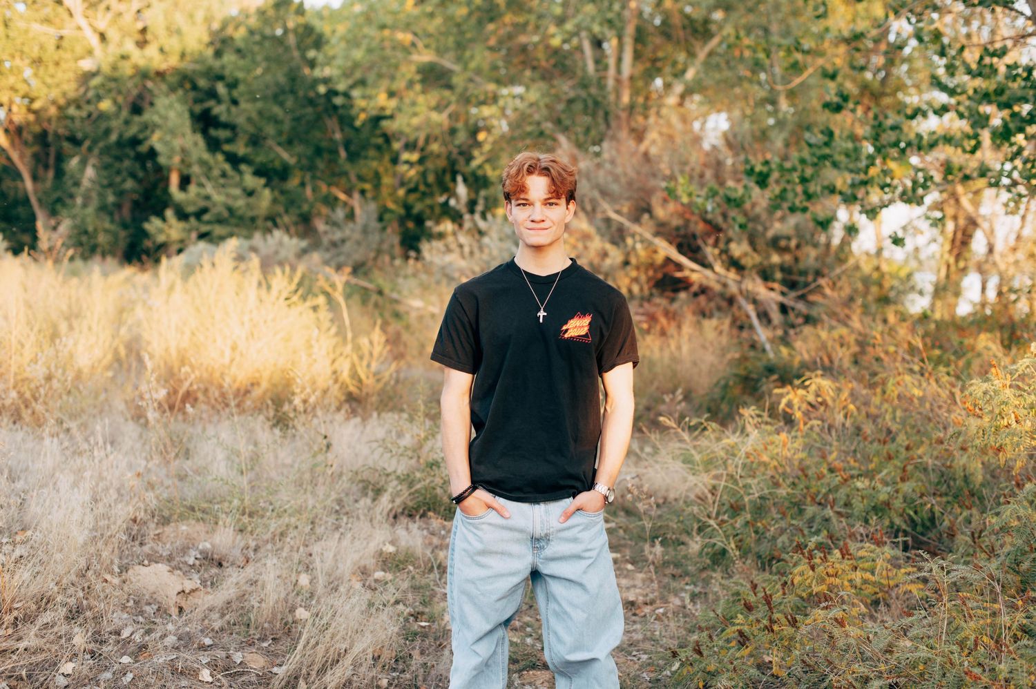 Young man in black t-shirt and jeans standing outdoors in a field with trees and dry grass in background.