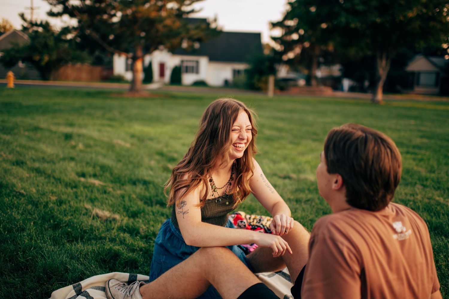 Two young people laughing and sharing snacks on a blanket in a green park at sunset.