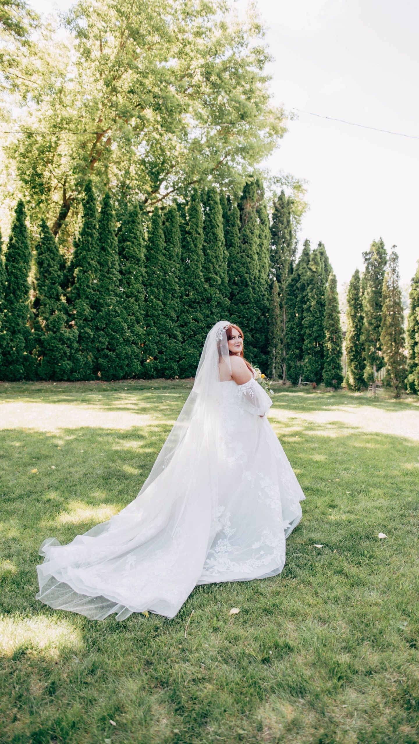 Bride in flowing white wedding dress twirling on grass with backdrop of evergreen trees.