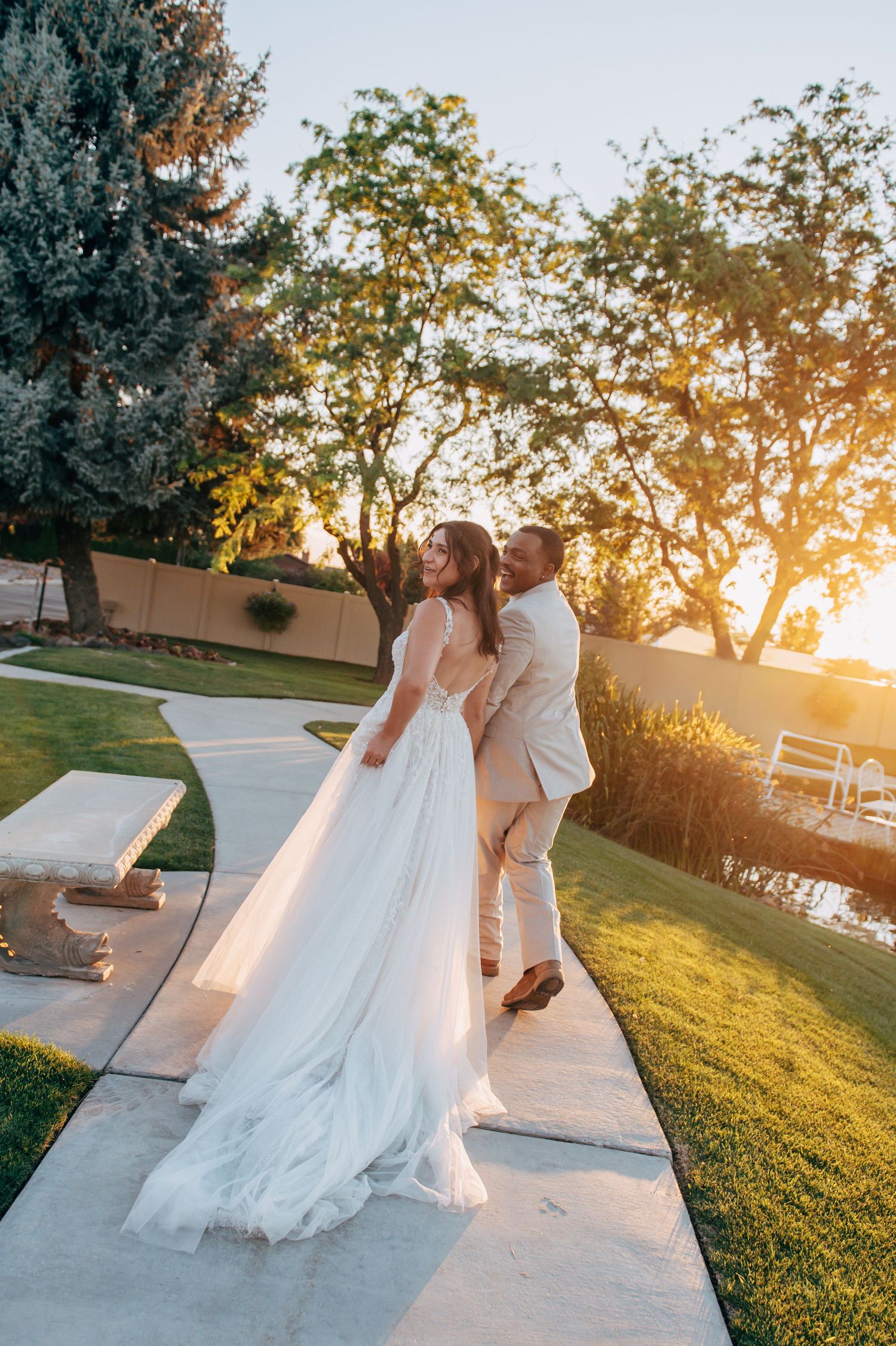 A couple shares a romantic embrace on a garden path at golden hour, bride in flowing white gown and groom in tan suit.