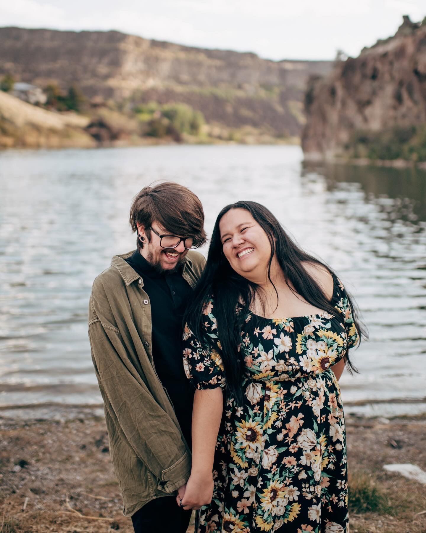 A smiling couple holds hands by a scenic lake with rocky cliffs, the woman in a floral dress laughing joyfully.