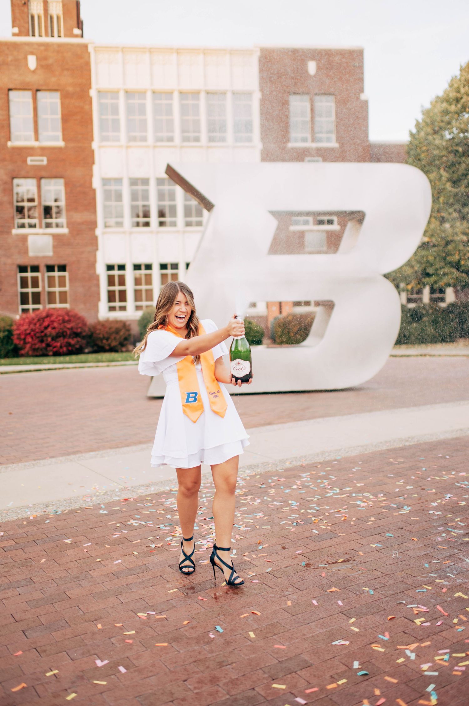 College graduate in white dress celebrates with champagne and confetti in front of large letter B on brick campus.